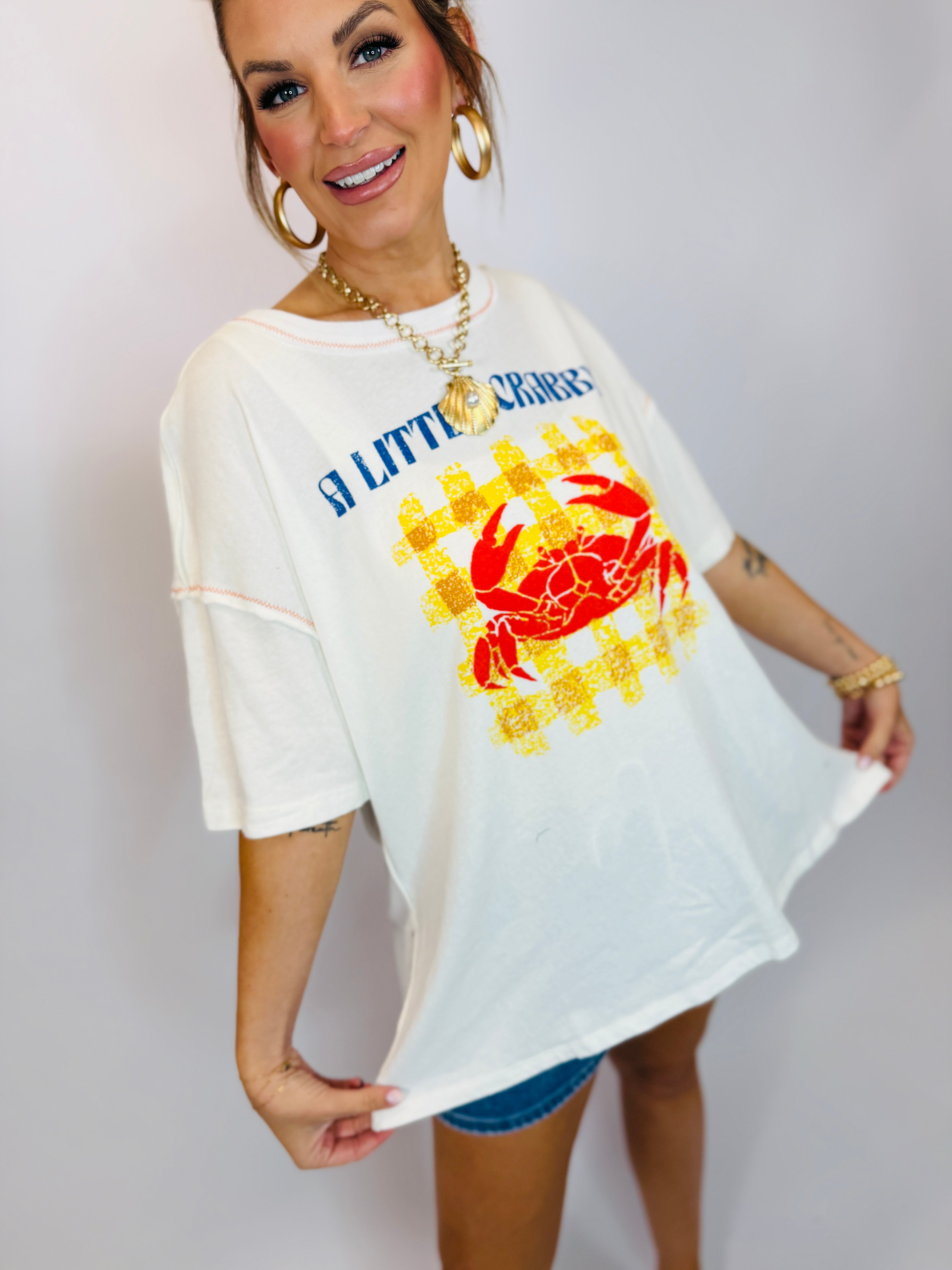 Woman wearing oversized white graphic t-shirt with colorful crab design and "A LITTLE CRABBY" text, styled with gold jewelry.