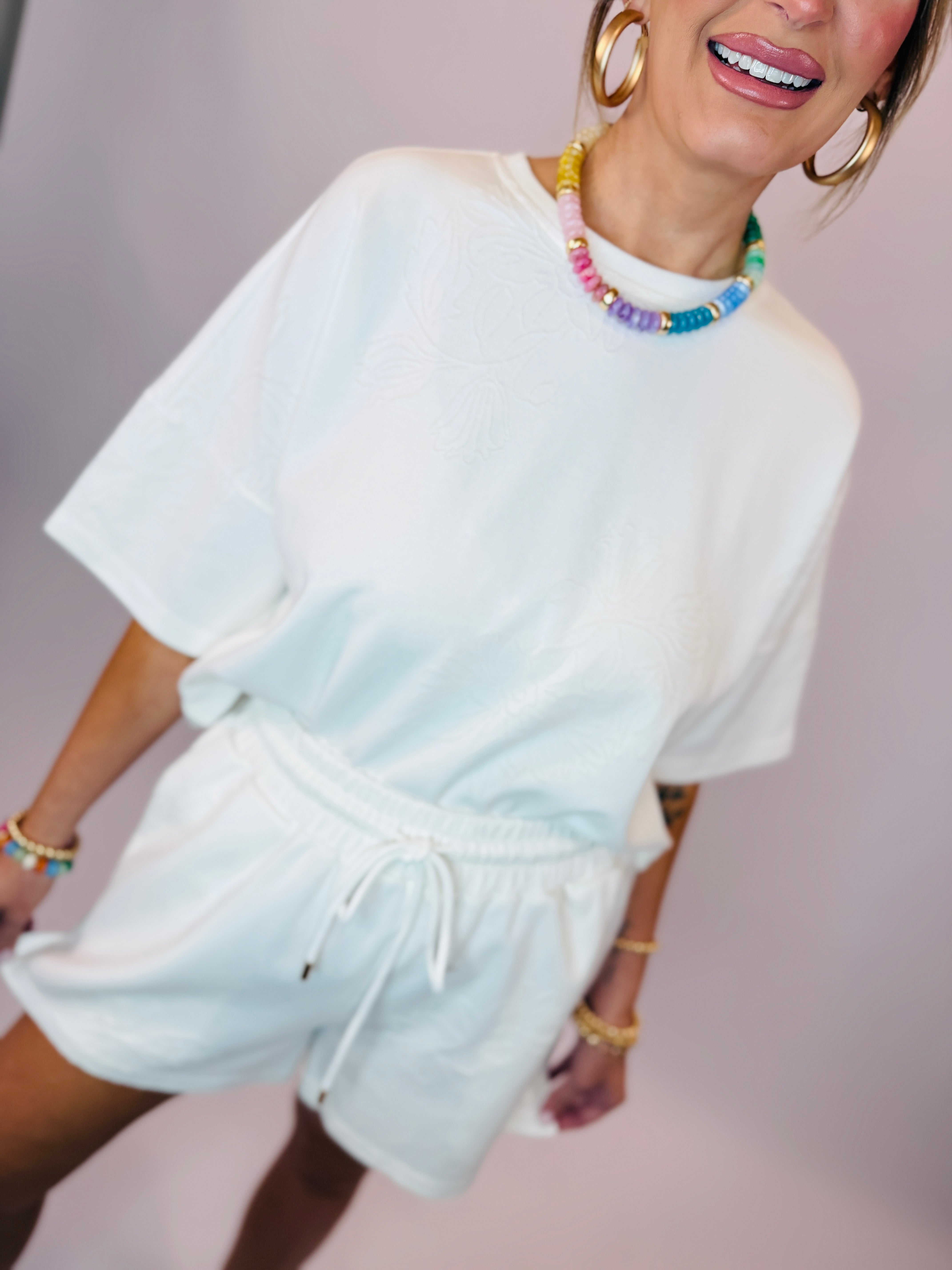 Woman wearing white oversized t-shirt and matching drawstring shorts with colorful beaded necklace and gold hoop earrings.