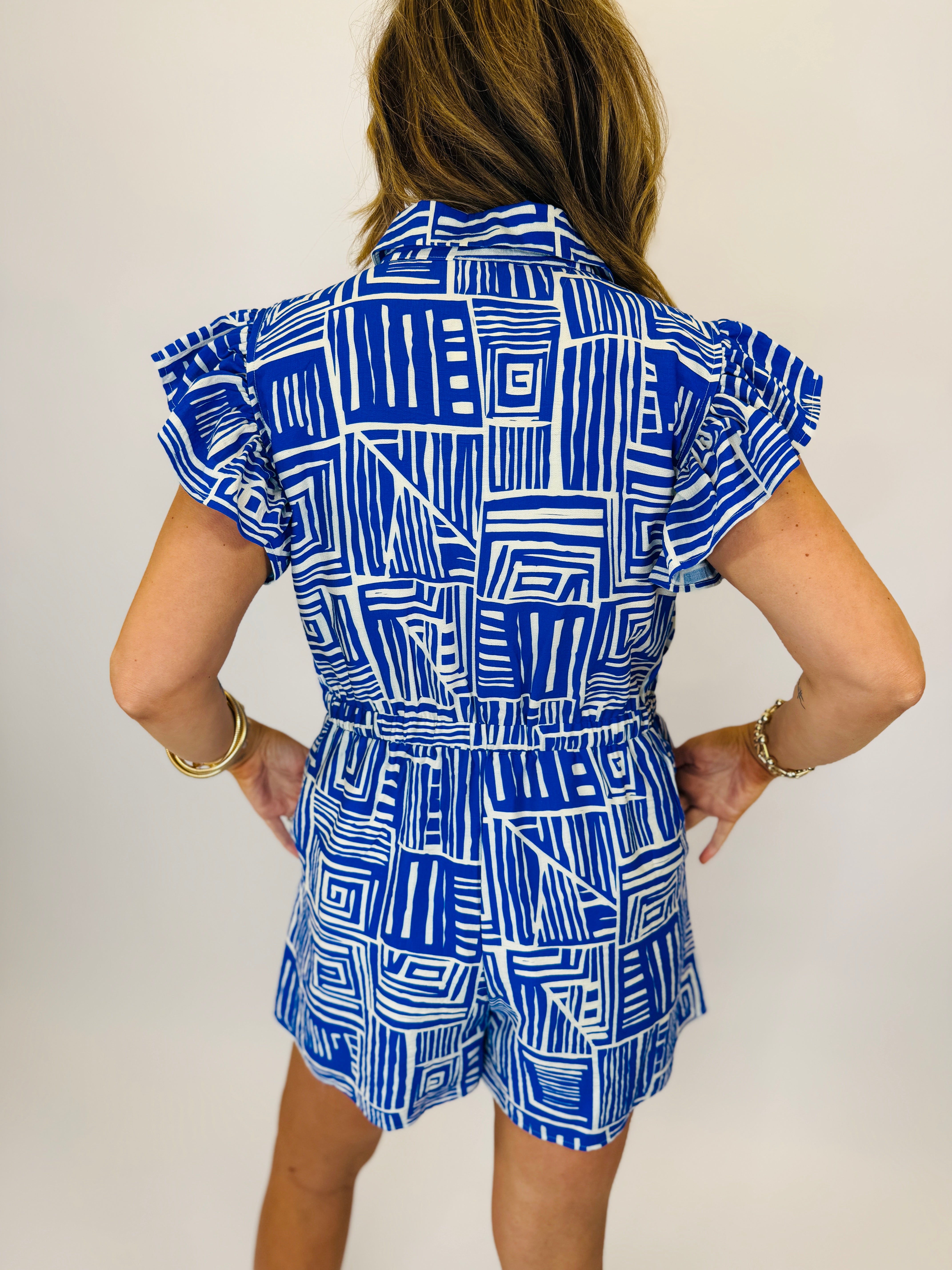 Woman wearing blue and white geometric patterned short-sleeve romper with tie waist belt, shown from behind.