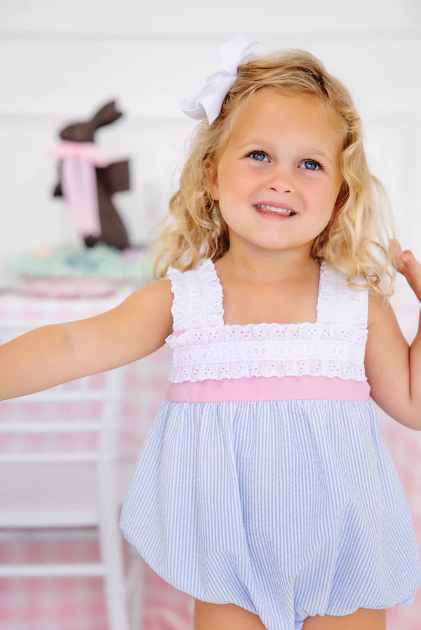 Young girl wearing white eyelet top with blue seersucker striped bubble romper and white hair bow.
