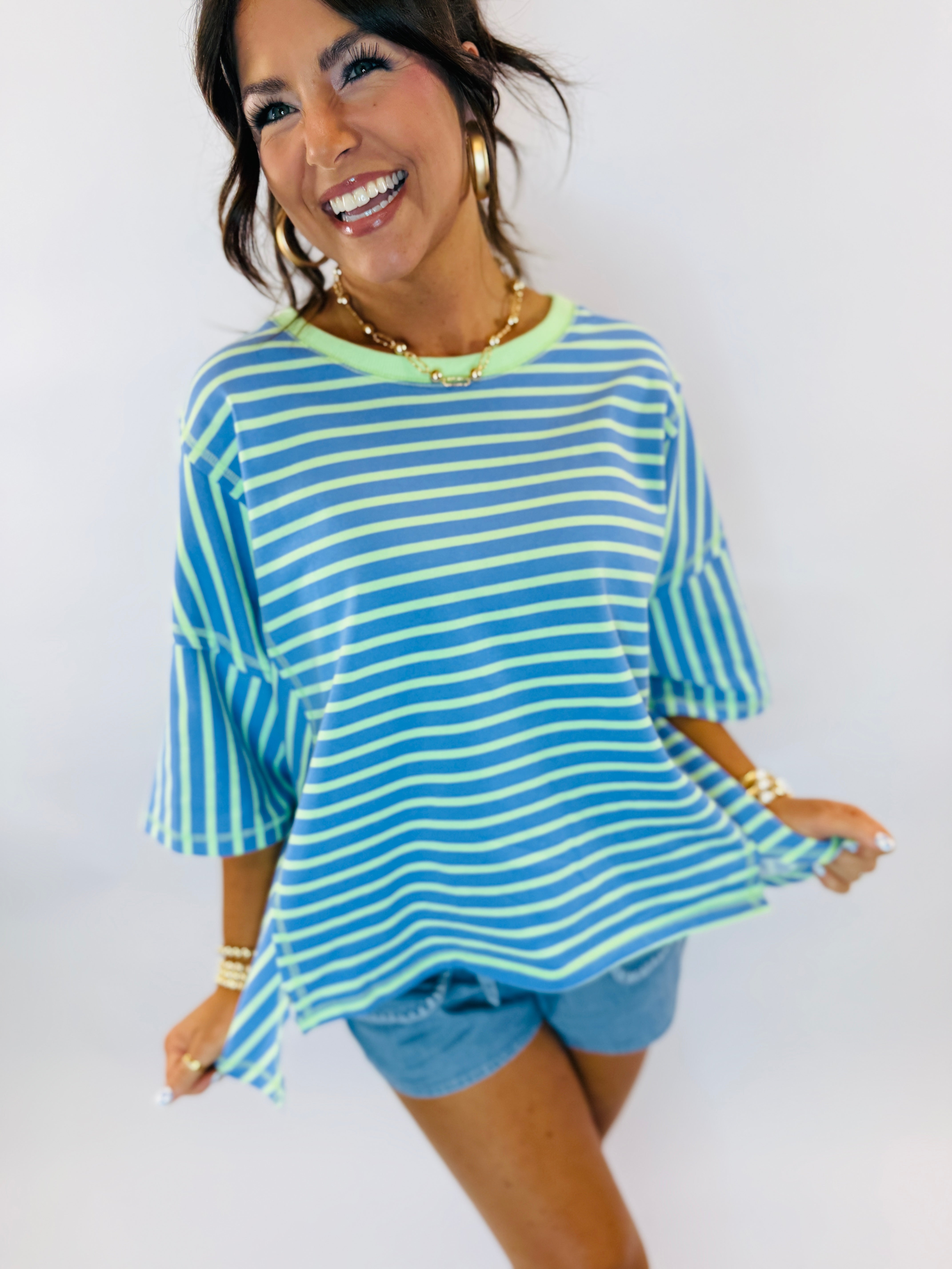 Woman wearing blue and green striped oversized t-shirt with matching blue shorts, smiling and accessorized with gold jewelry.