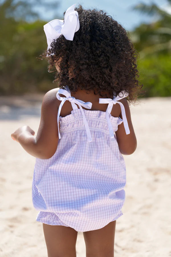 Baby girl wearing lavender gingham Rosey Romper with white bow ties and matching hair bow on beach