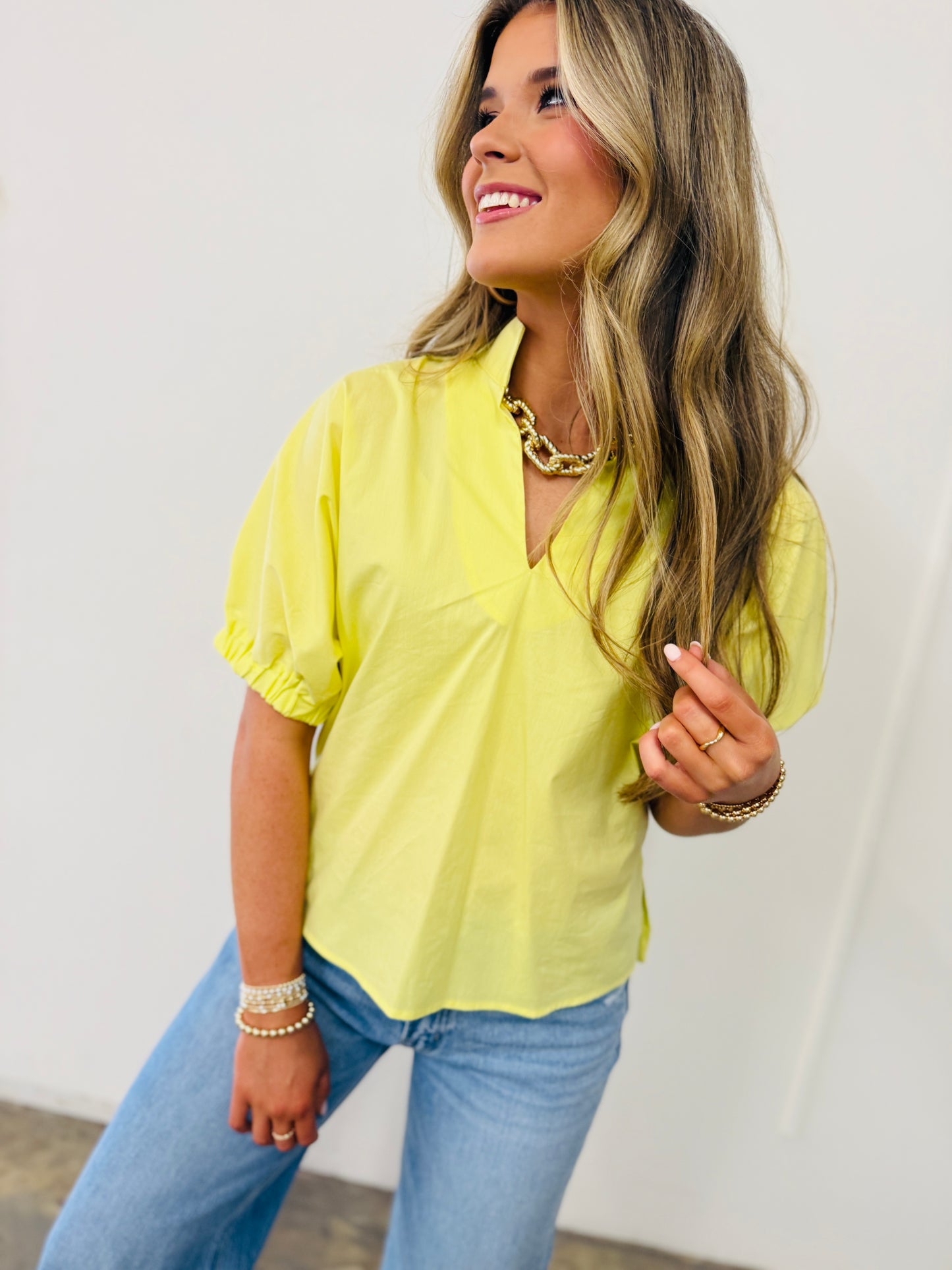 Woman wearing yellow Martha Soft Top with short sleeves and v-neck, paired with blue jeans and gold jewelry.