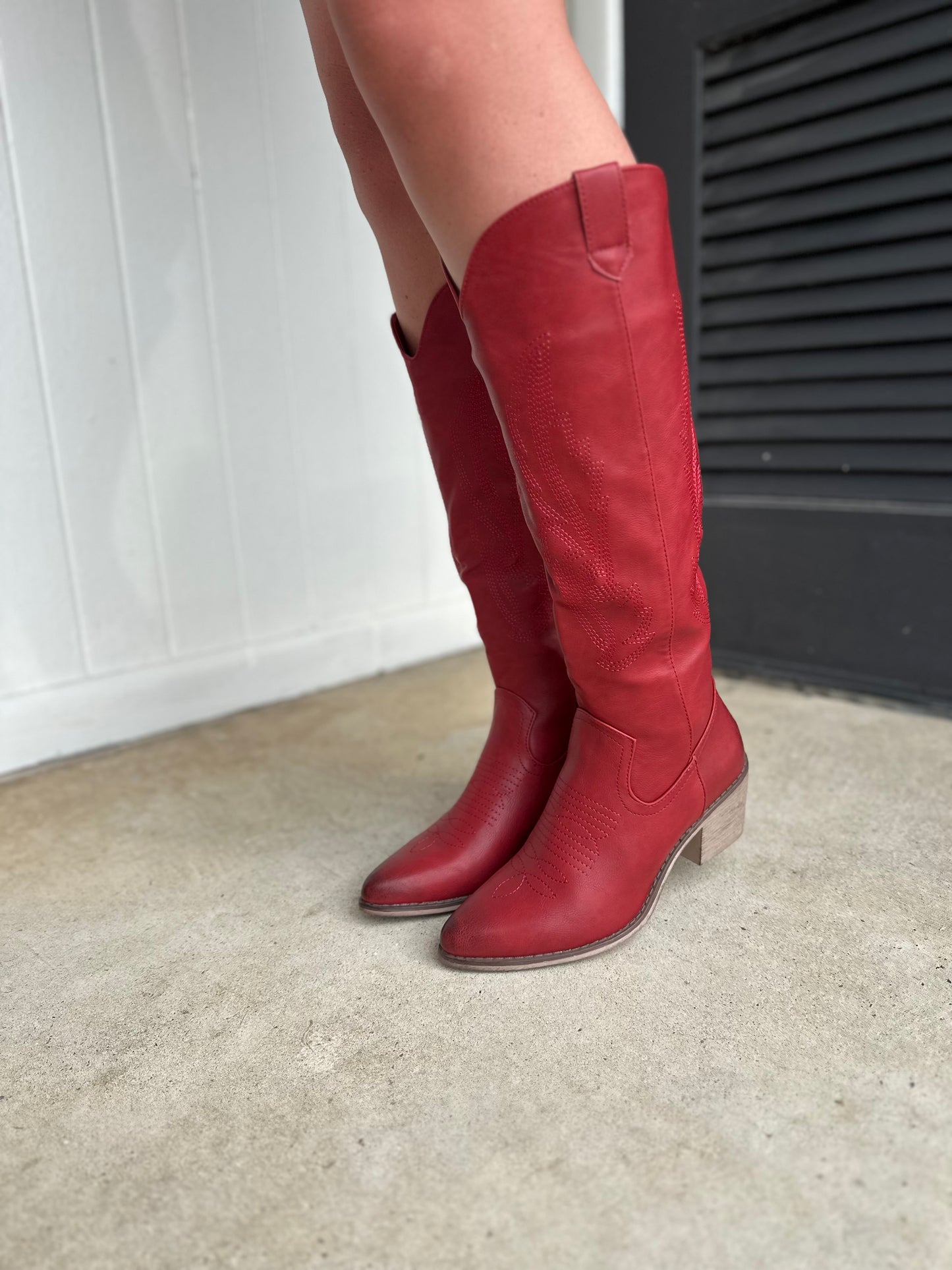 Red leather knee-high cowboy boots with decorative stitching and block heels worn on concrete surface.