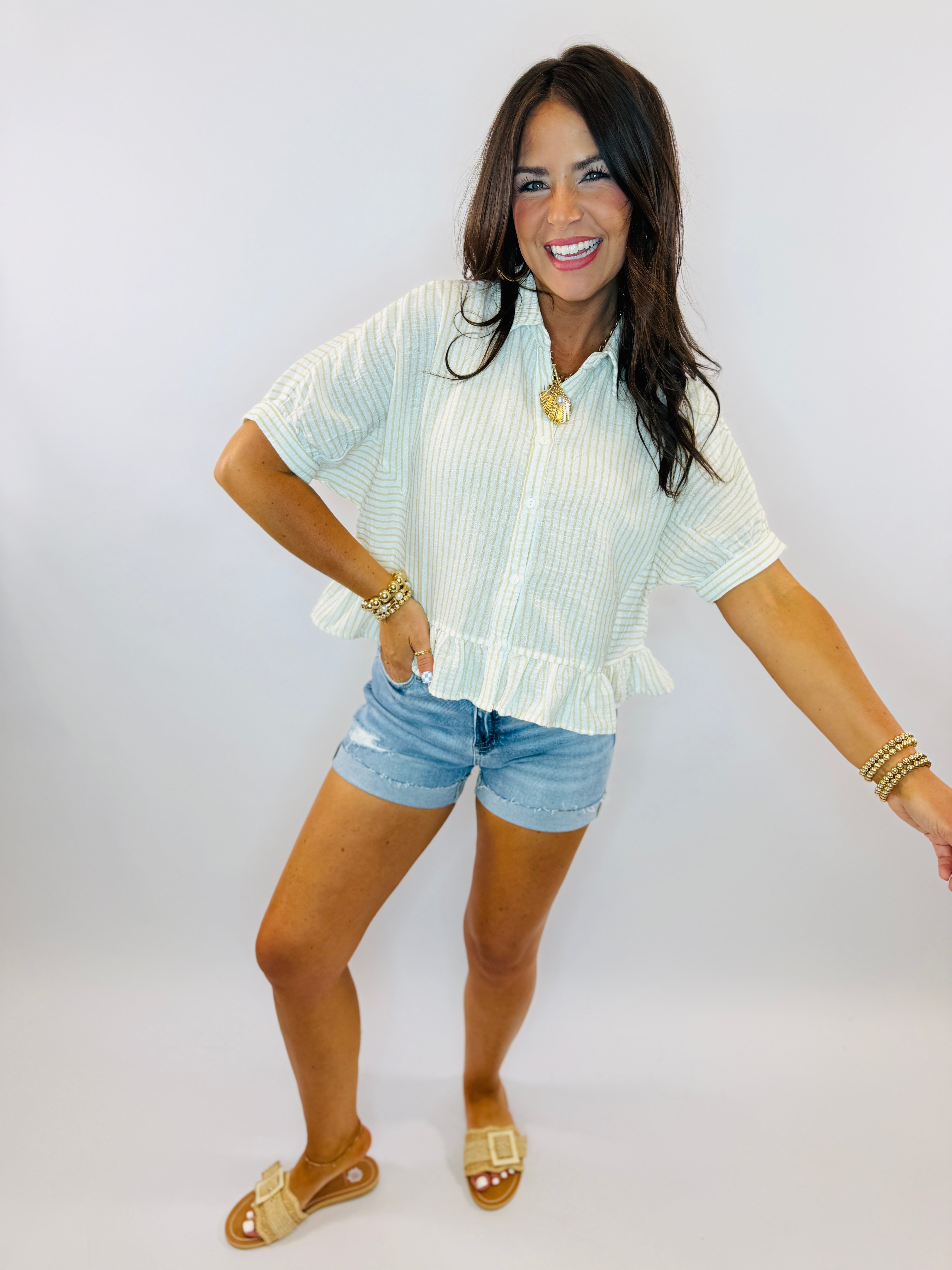 Woman wearing light blue striped button-up top with ruffle hem, denim shorts, and gold jewelry accessories.