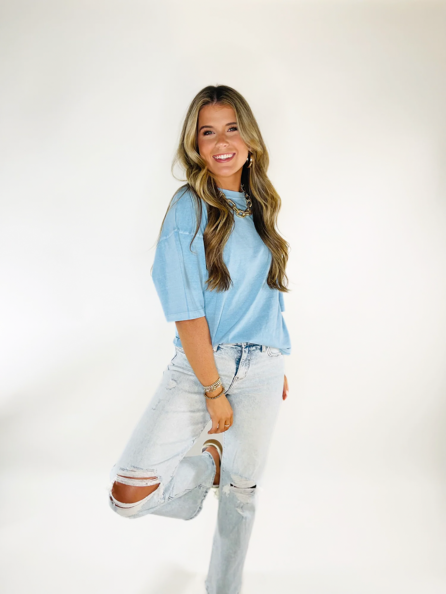 Woman wearing light blue oversized t-shirt with ripped jeans and layered necklaces against white background