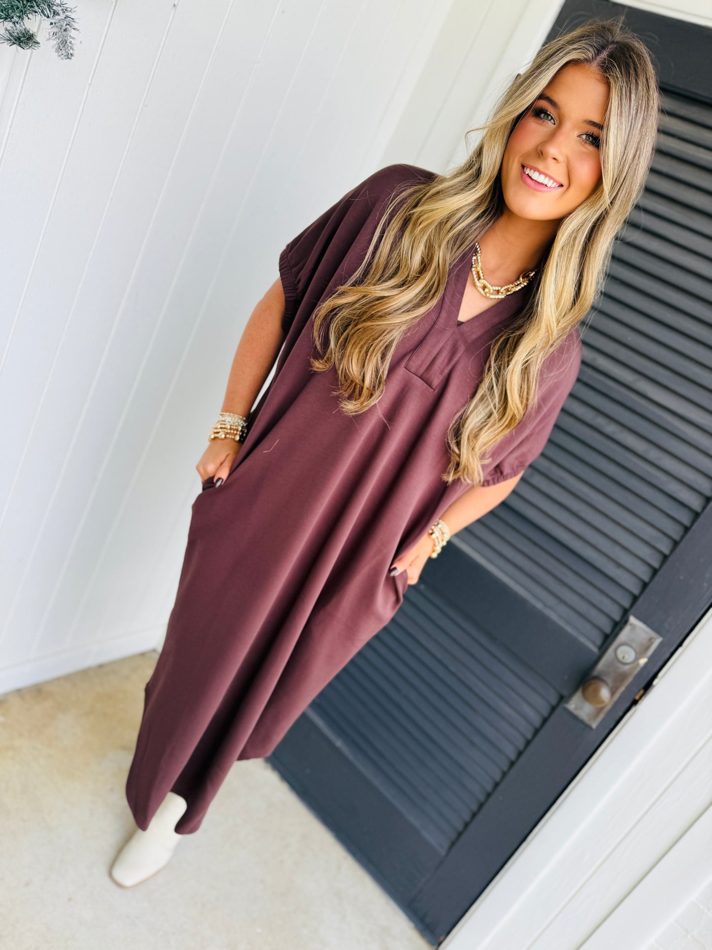Woman wearing burgundy flowy midi dress with short sleeves, styled with gold jewelry and white sneakers.
