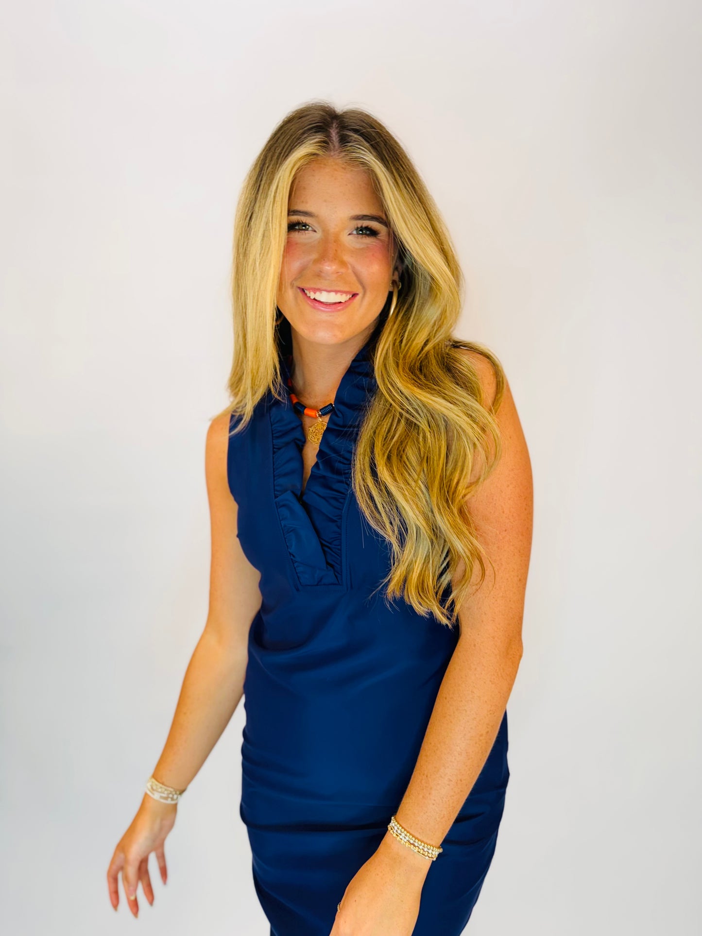 Stella Dress in navy blue sleeveless ruffle V-neck style, worn by smiling blonde model with gold bracelets.