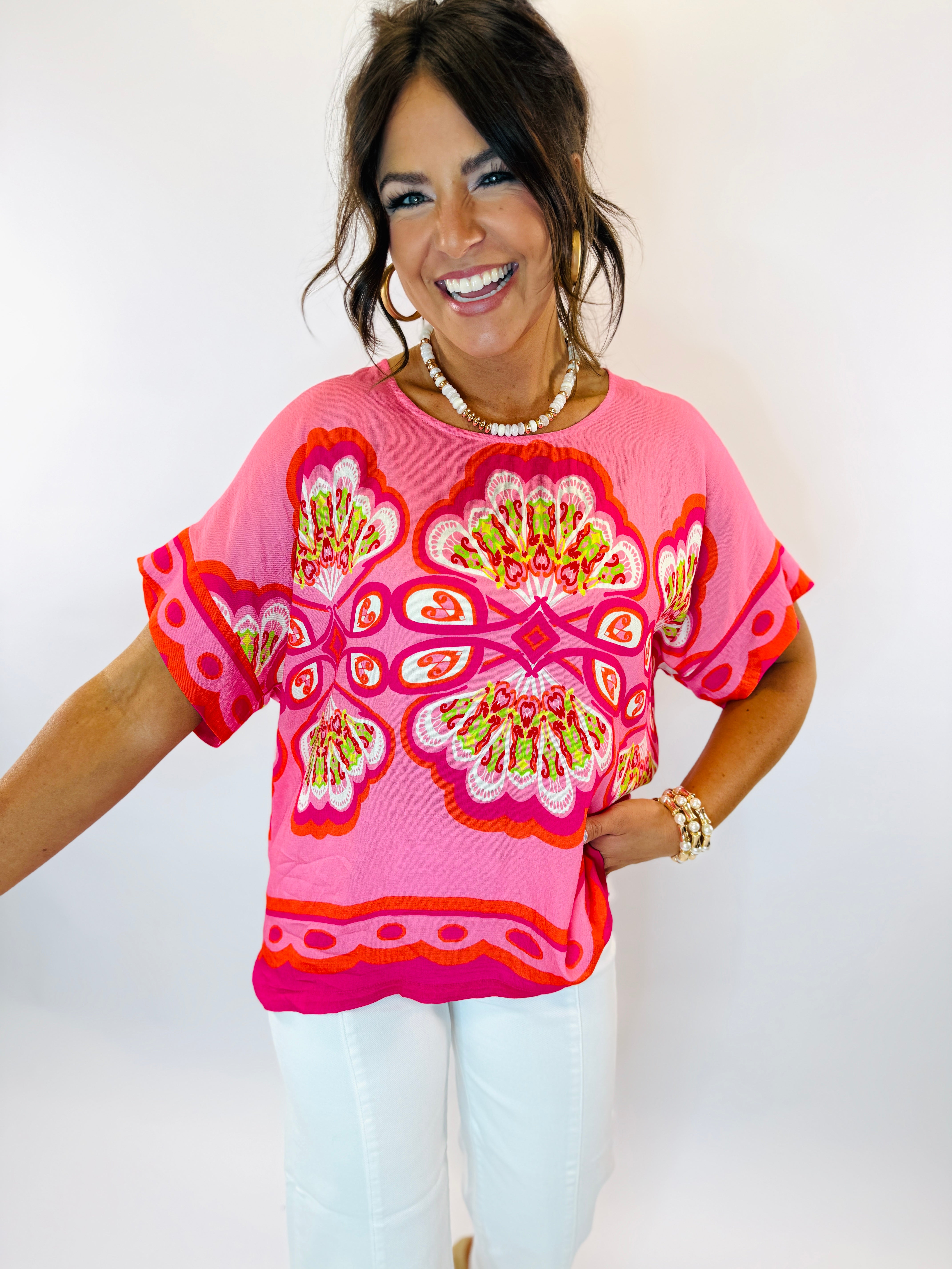 Woman wearing bright pink Laci top with colorful floral pattern and white jeans, smiling at camera.