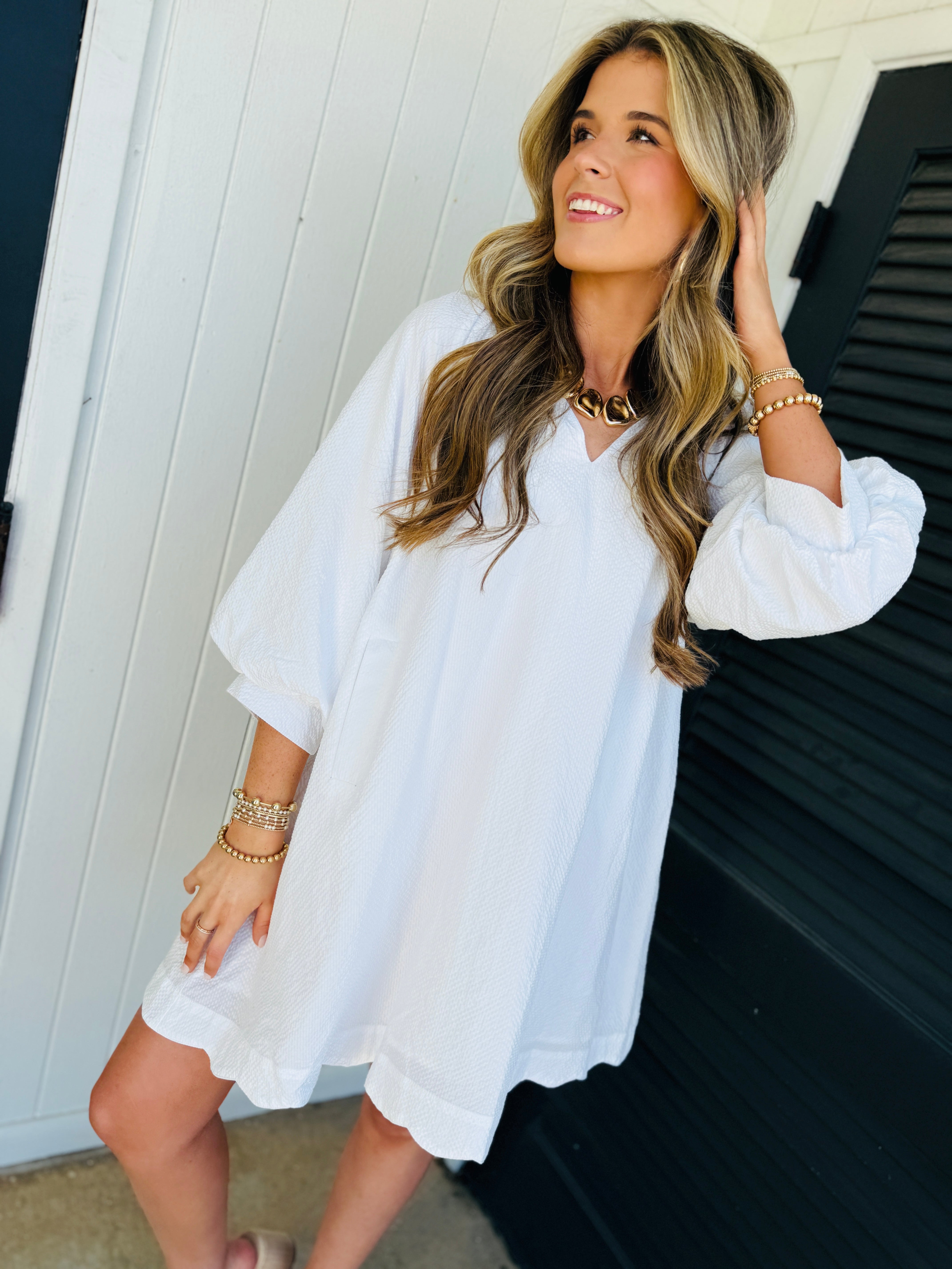 Woman wearing white long-sleeved Libby Maple Classic Dress with gold jewelry, standing against white wooden backdrop.