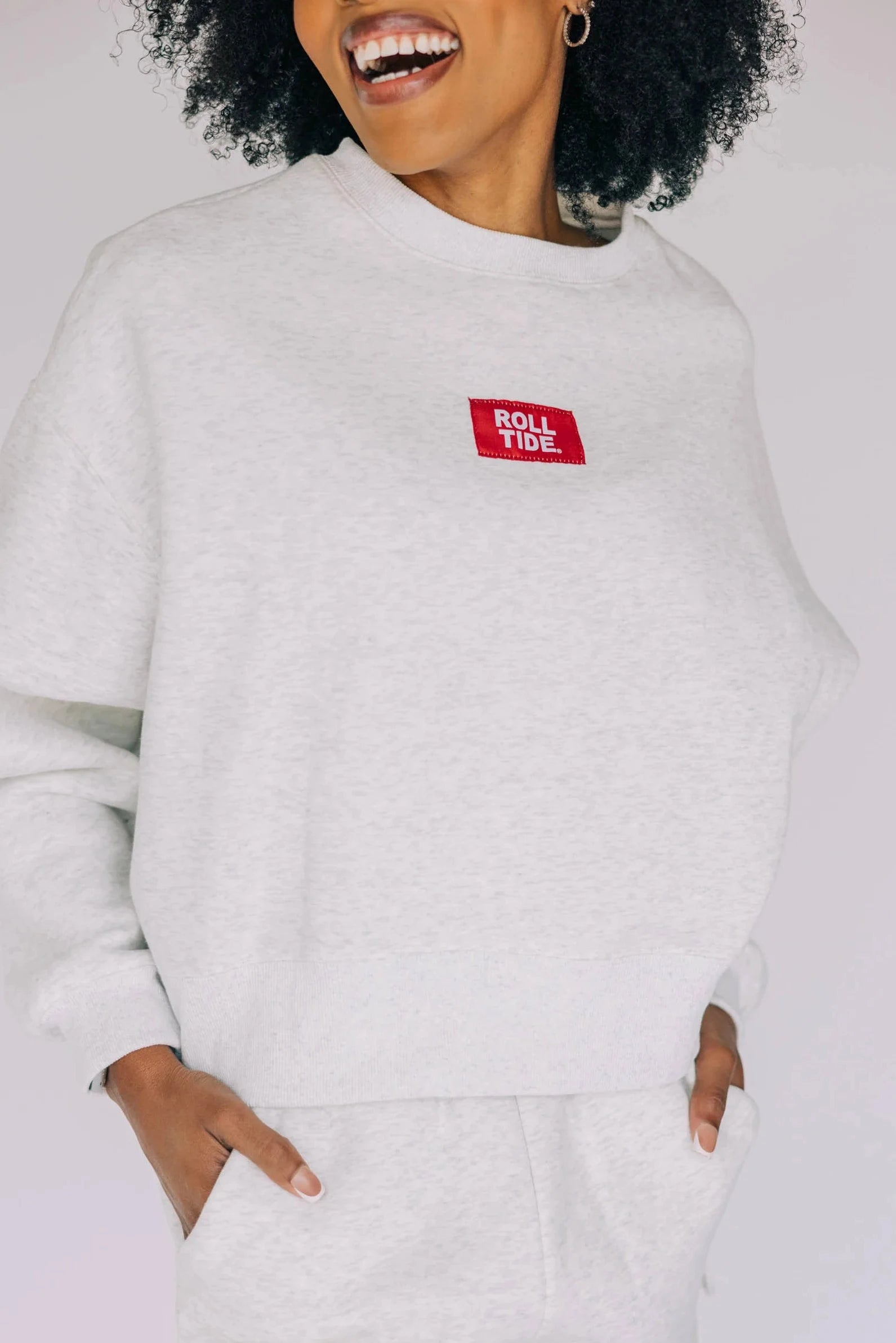 Woman wearing a light heather gray boxy crewneck sweatshirt with a red "Roll Tide" patch logo.