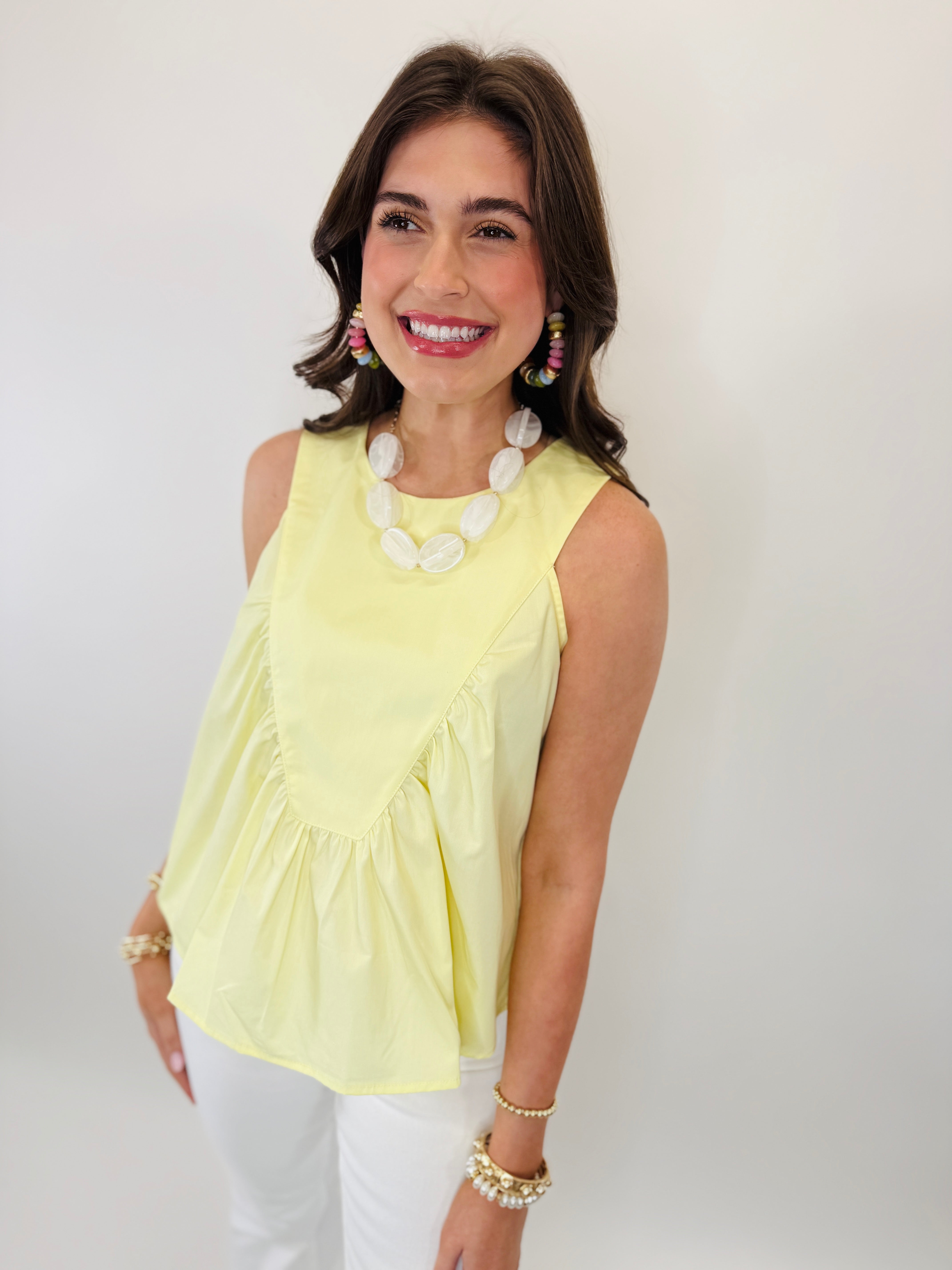 Woman wearing yellow sleeveless Fran top with white pearl necklace and colorful earrings, smiling at camera.