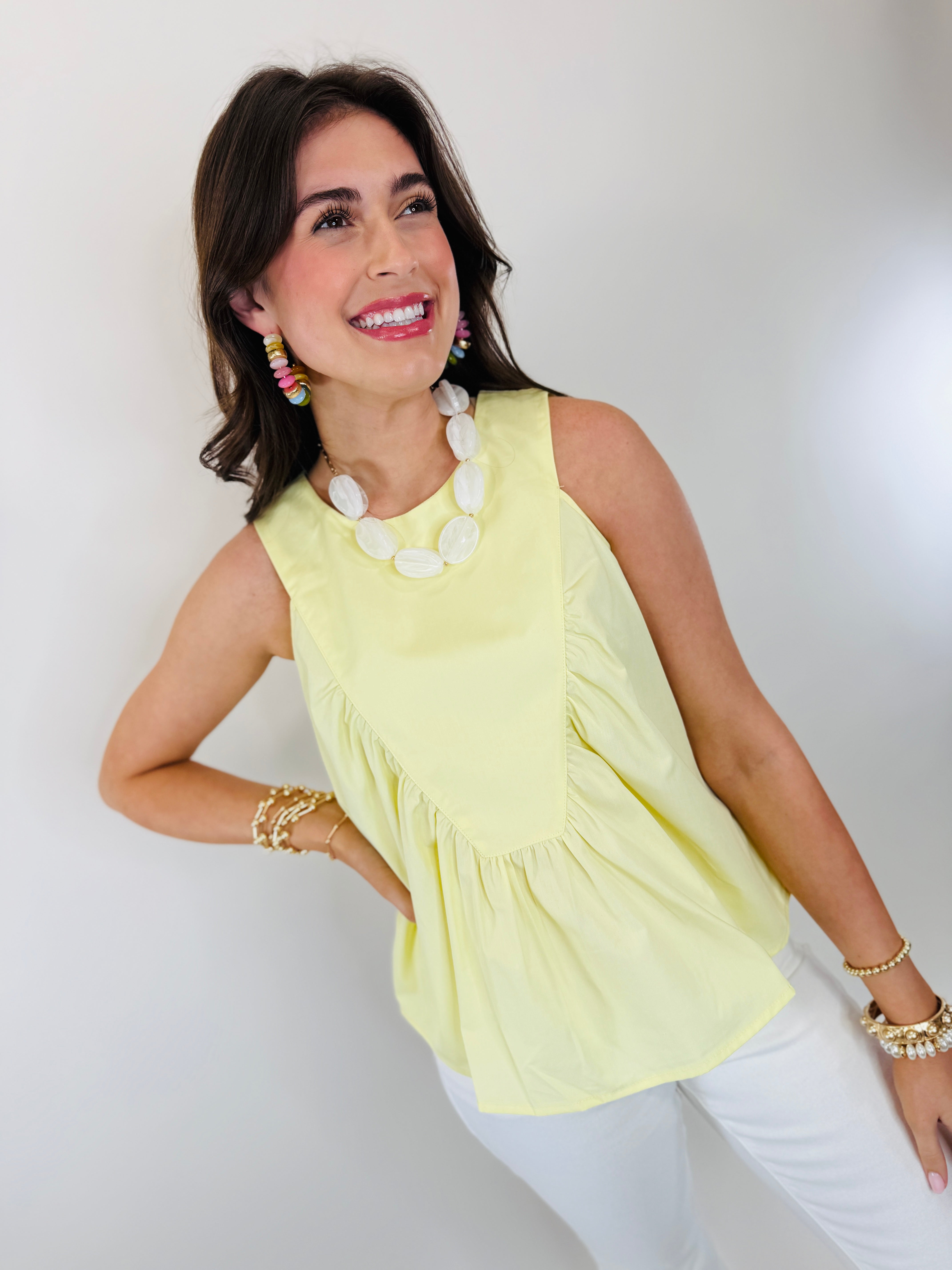 Woman wearing yellow sleeveless ruffle top with white statement necklace and colorful earrings, smiling at camera.