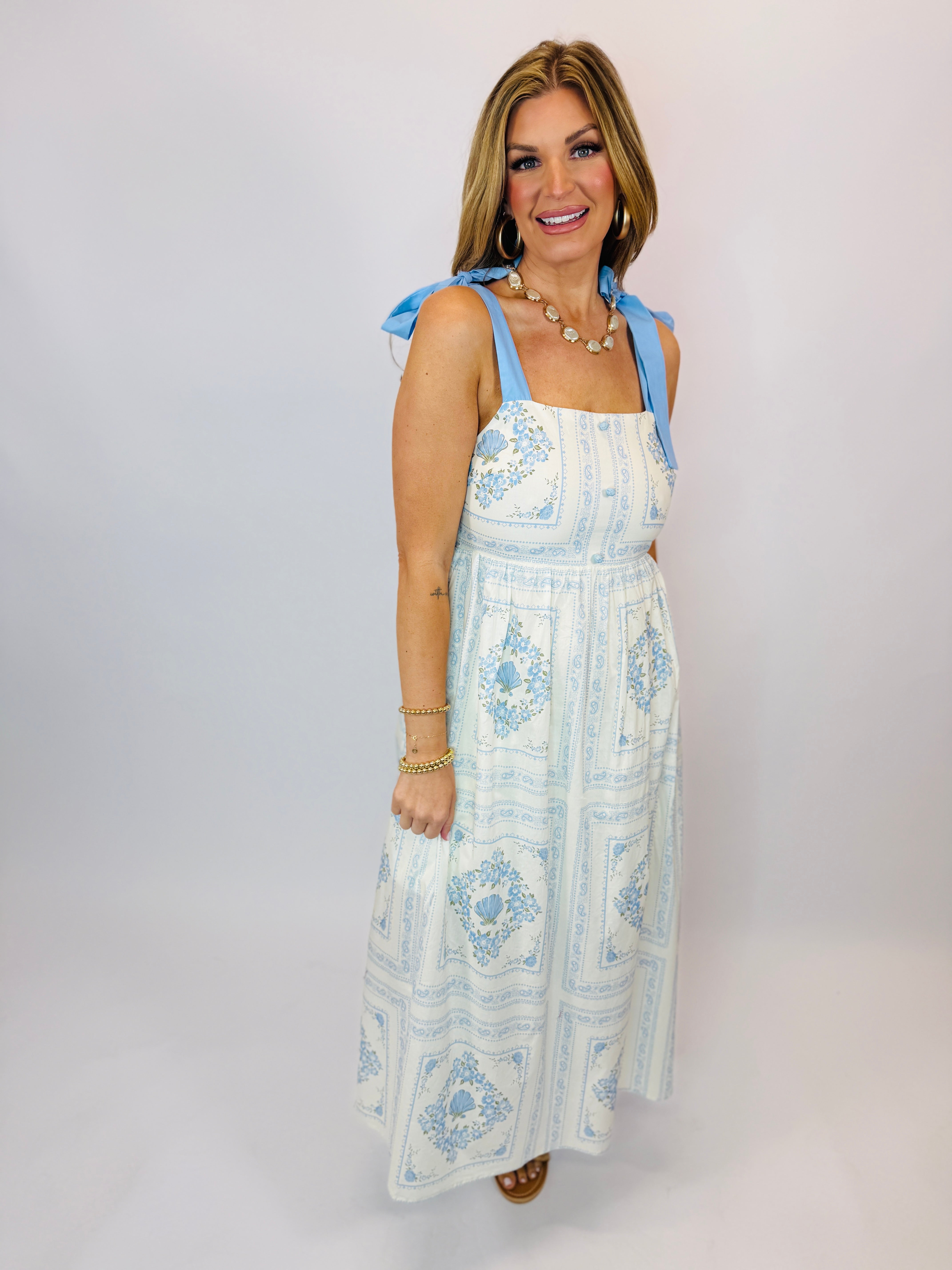 Woman wearing white and blue patterned maxi dress with tie shoulder straps and decorative medallion print.
