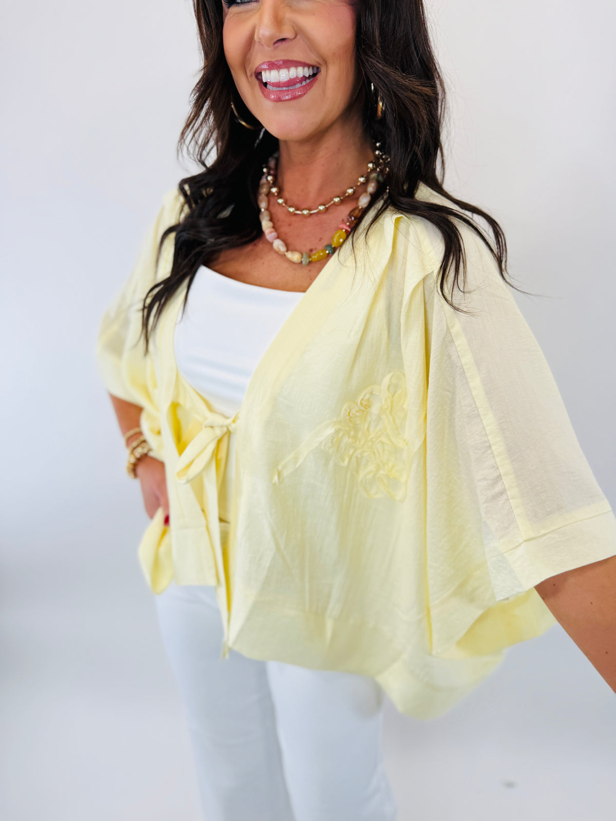 Woman wearing yellow sheer kimono over white top with pearl necklace, smiling against white background.