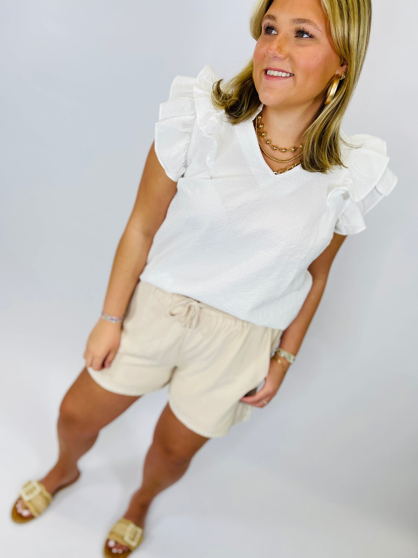 White ruffle sleeve top with dramatic shoulder details worn with beige shorts and gold accessories