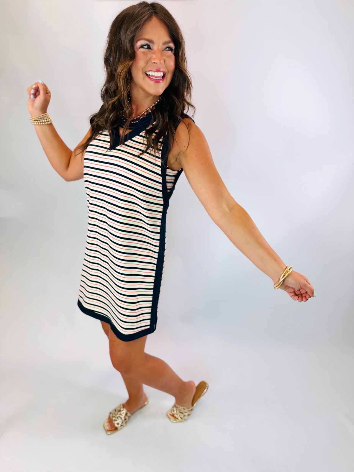 Woman wearing the Jordan Dress, a sleeveless navy-trimmed striped shift dress with coral and white stripes, styled with go...