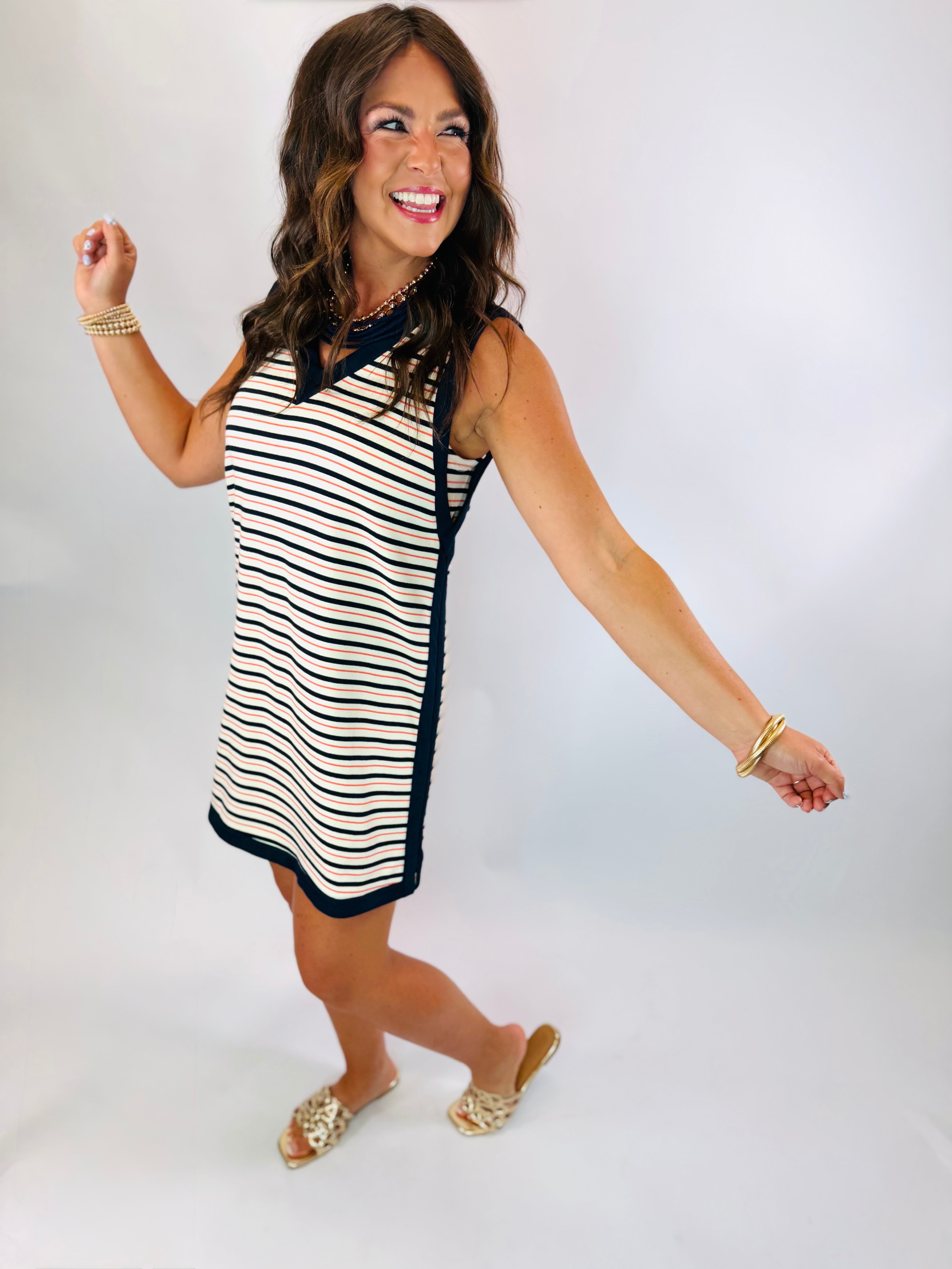 Woman wearing the Jordan Dress, a sleeveless navy-trimmed striped shift dress with coral and white stripes, styled with go...