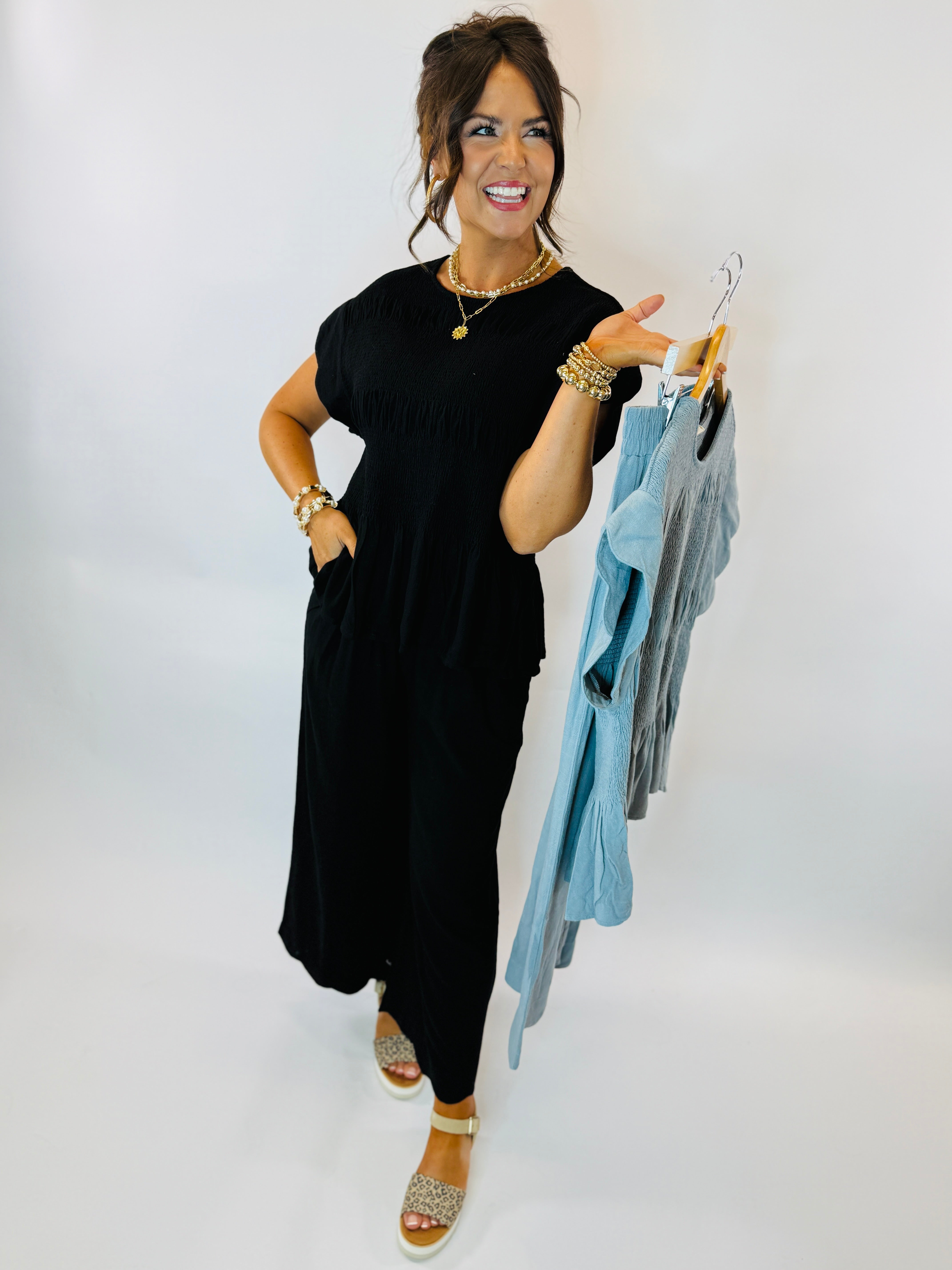 Woman wearing black Marlee top and pants set, holding blue clothing items on hangers while smiling.