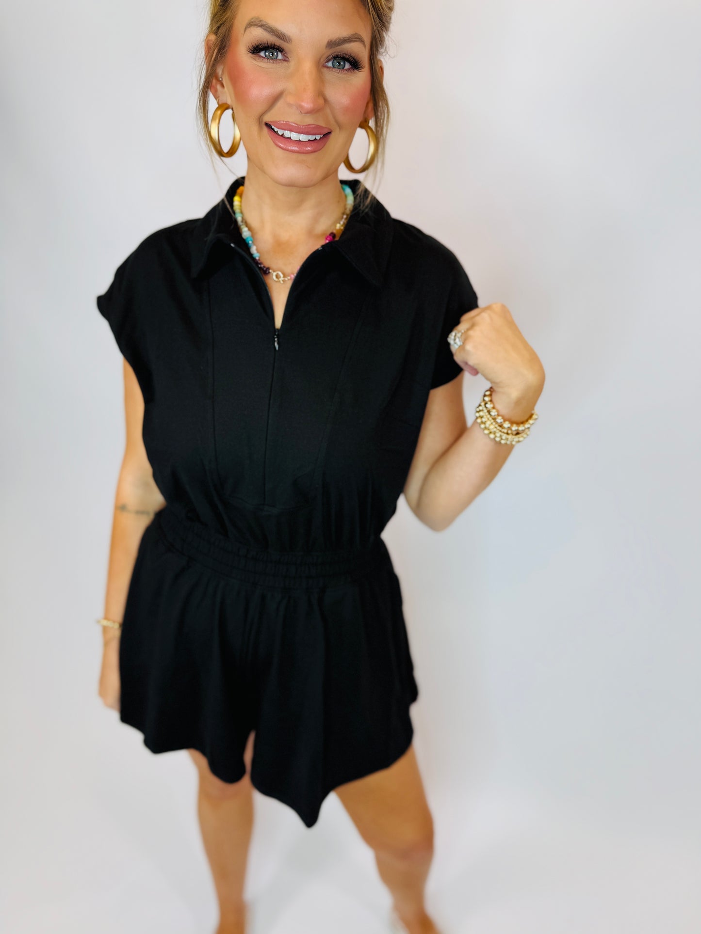 Woman wearing black short-sleeve collared romper with tie waist, styled with gold jewelry and hoop earrings.