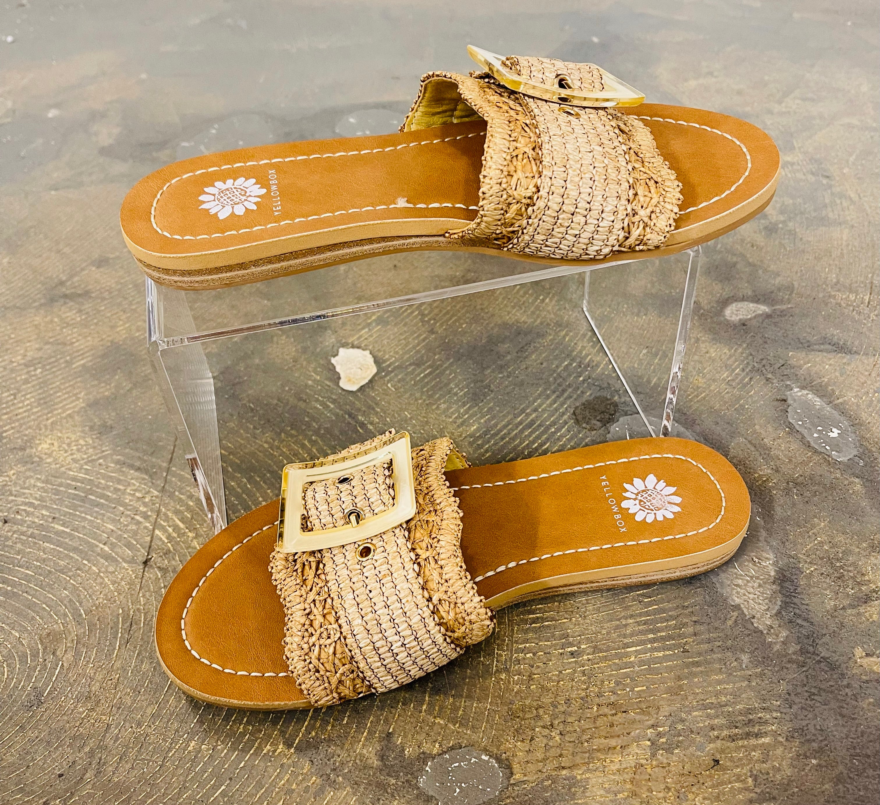 Two woven straw sandals with buckle straps and orange soles displayed on clear acrylic stand at beach