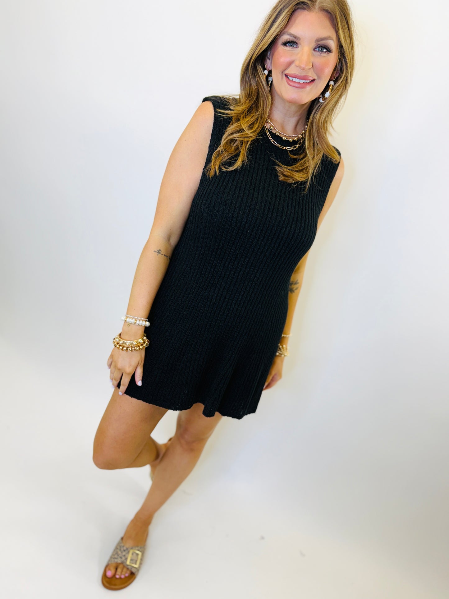 Woman wearing black sleeveless ribbed mini dress with gold jewelry and sandals, smiling at camera.