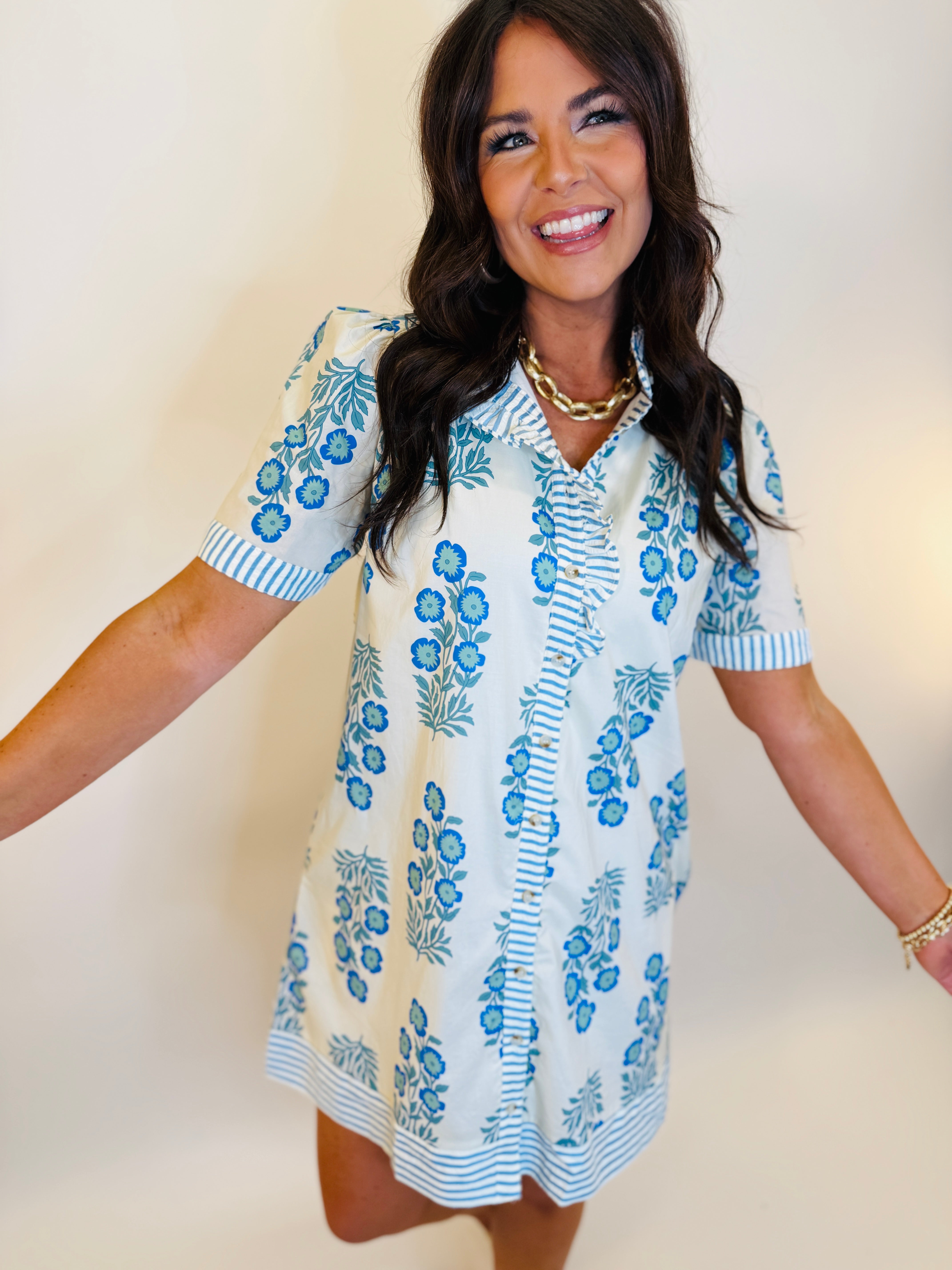 Woman wearing white short-sleeve dress with blue floral pattern and striped trim details, smiling against neutral background.