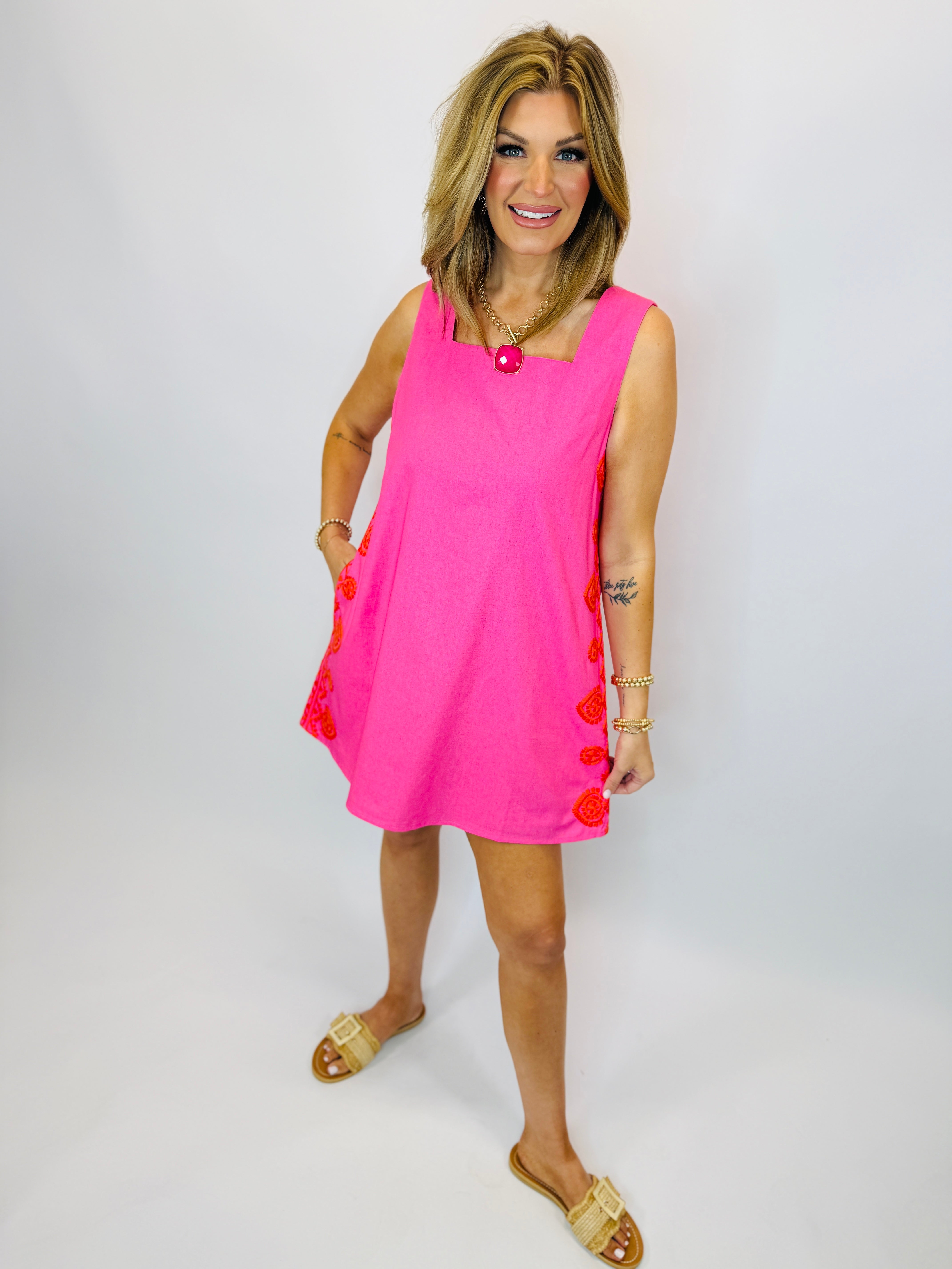 Woman wearing bright pink sleeveless Marlene dress with red pom-pom trim and gold sandals