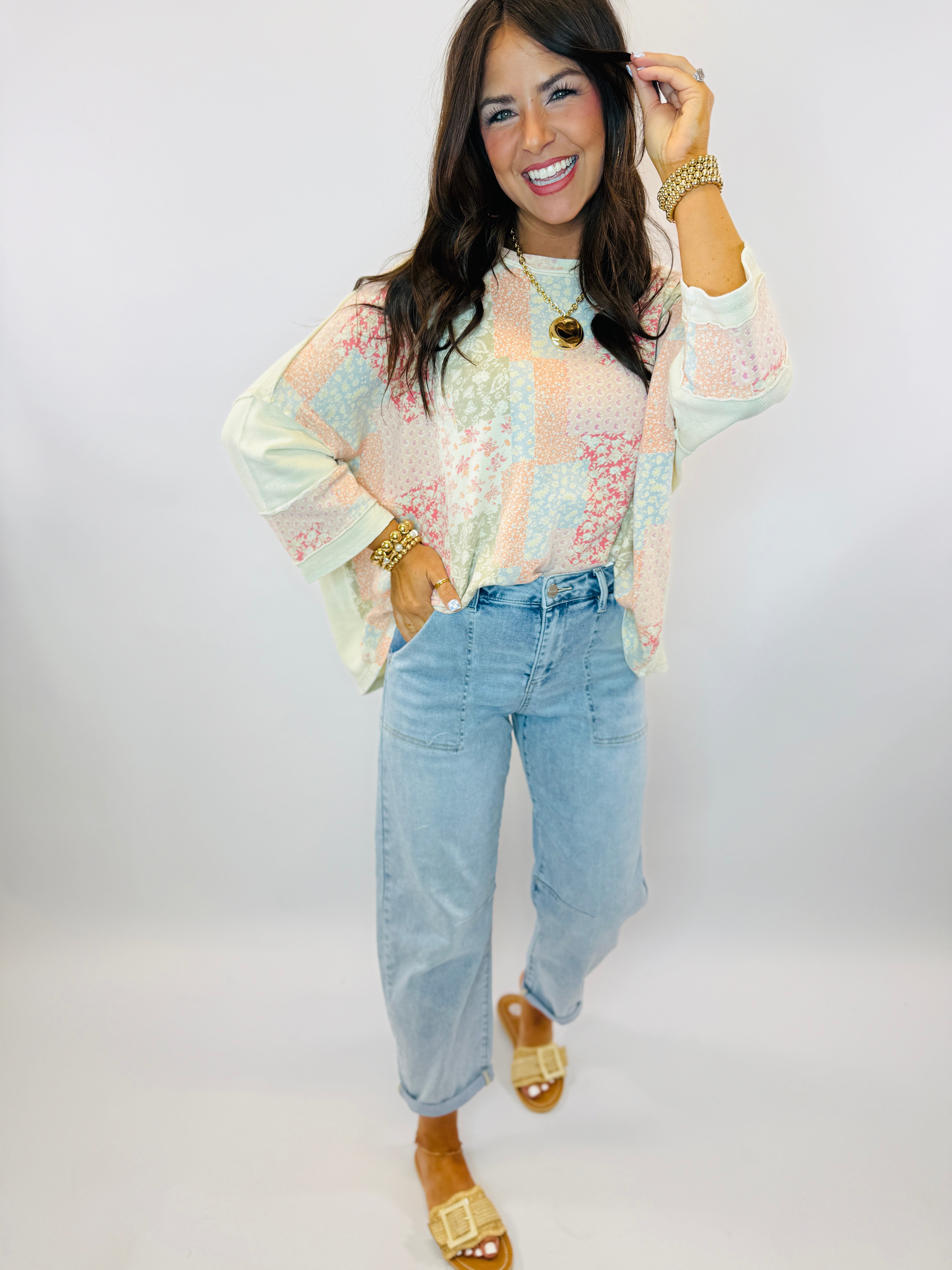 Woman wearing pastel patchwork long-sleeve top with light blue jeans and gold sandals, smiling and posing.