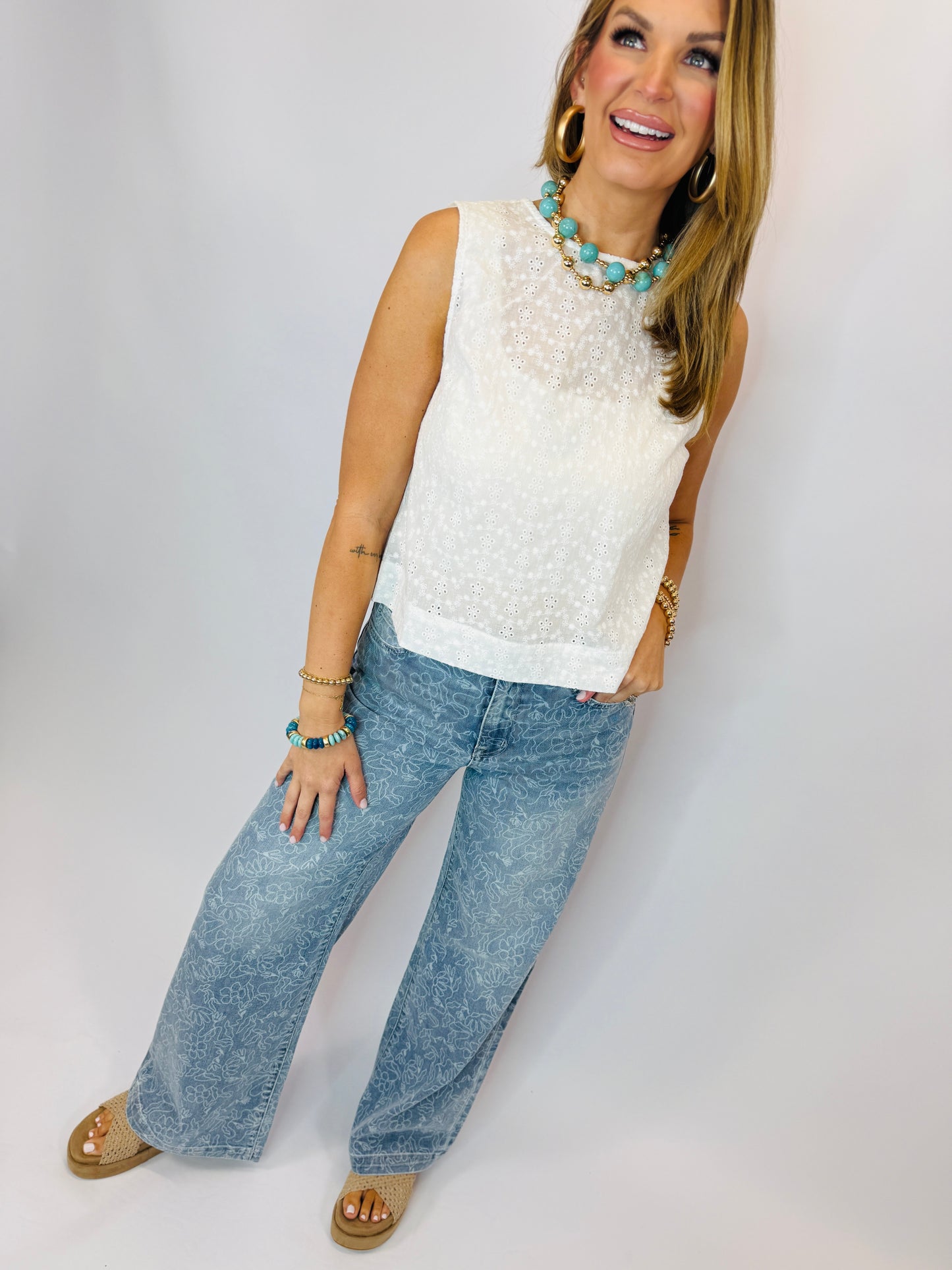 Woman wearing white eyelet top and blue floral patterned straight-leg jeans with turquoise jewelry and platform sandals