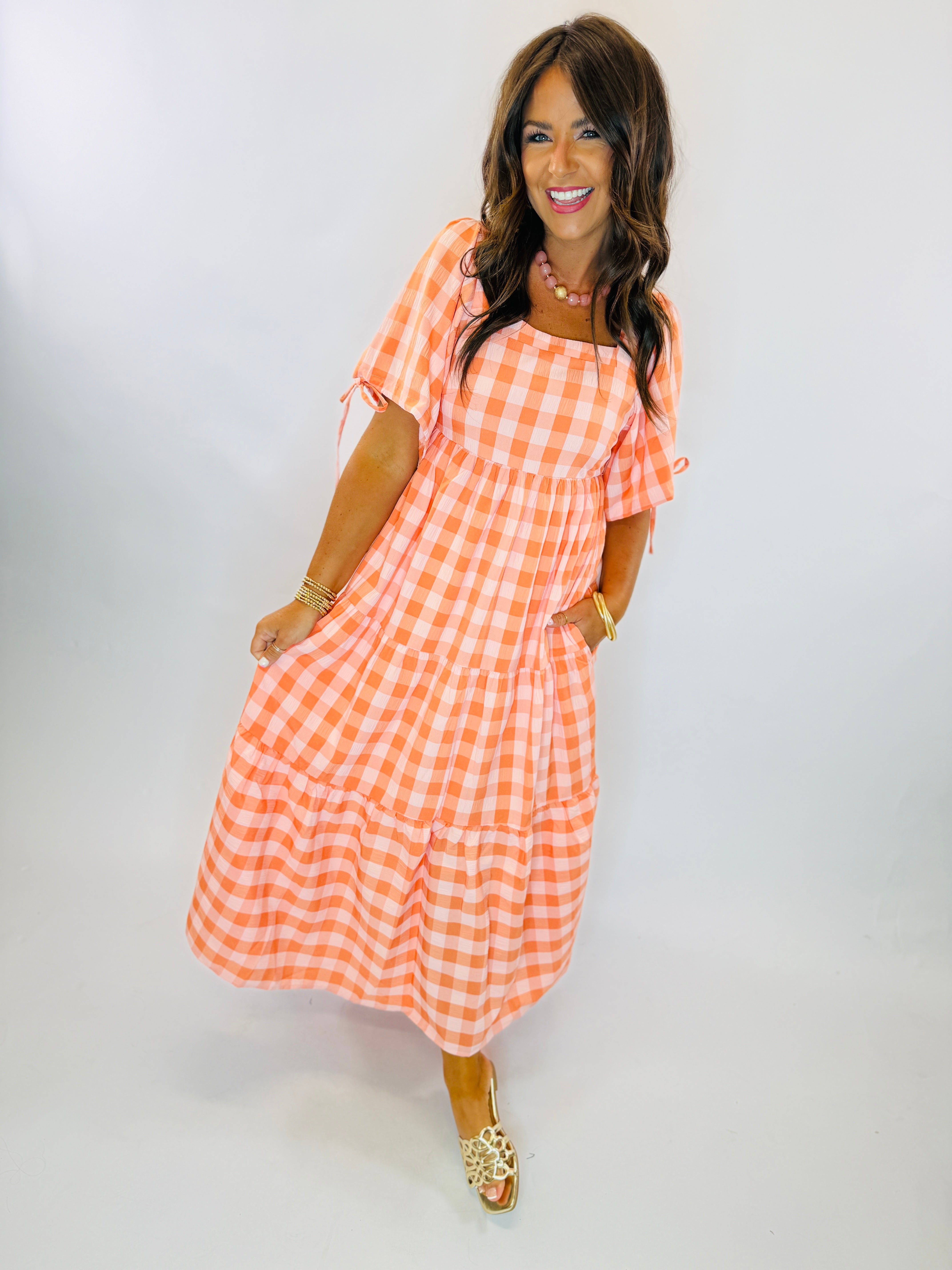 Woman wearing orange and white gingham tiered maxi dress with flutter sleeves and tie details.