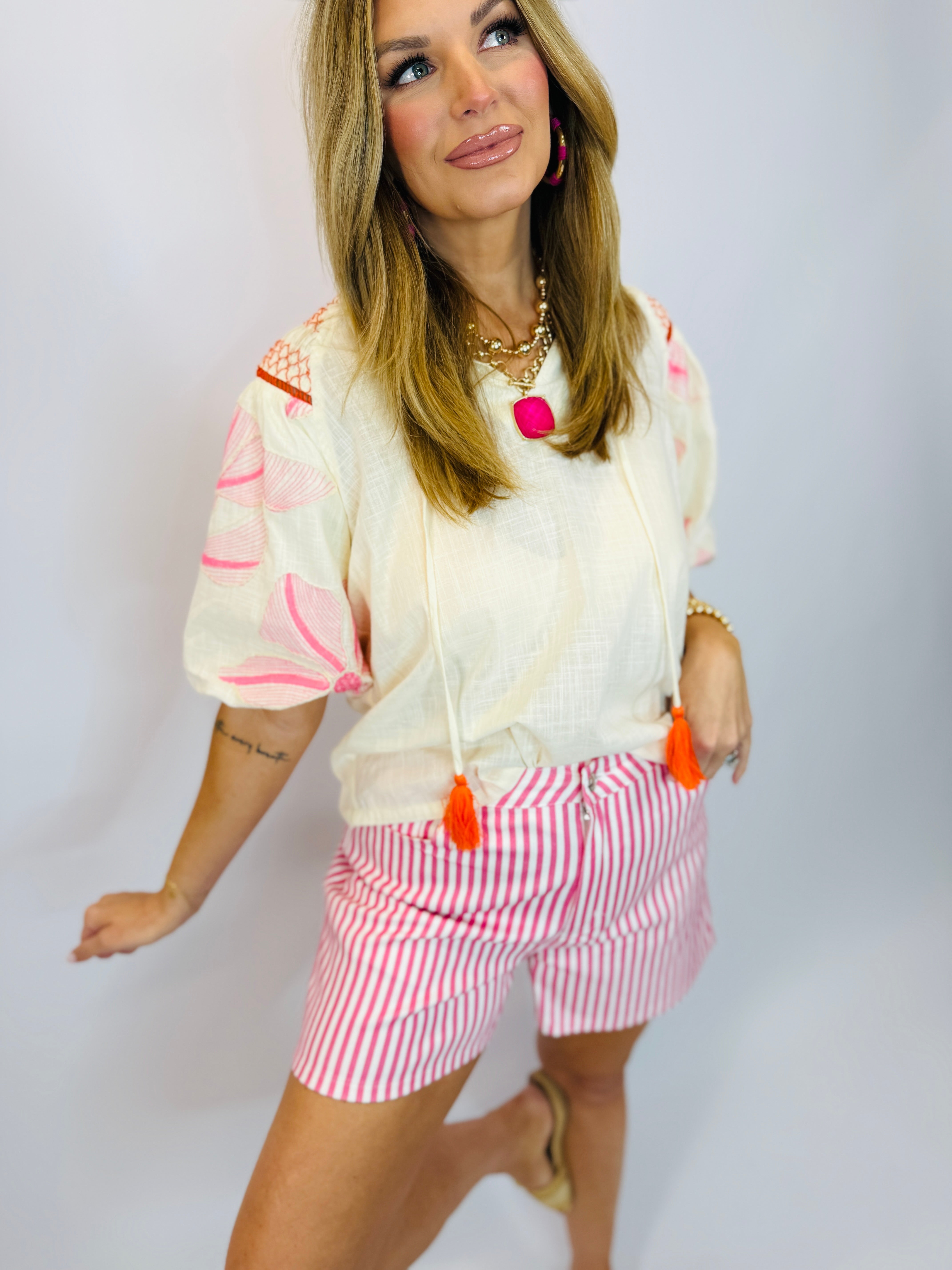 Woman wearing white embroidered Vera top with pink floral details and orange tassel ties, paired with striped shorts.
