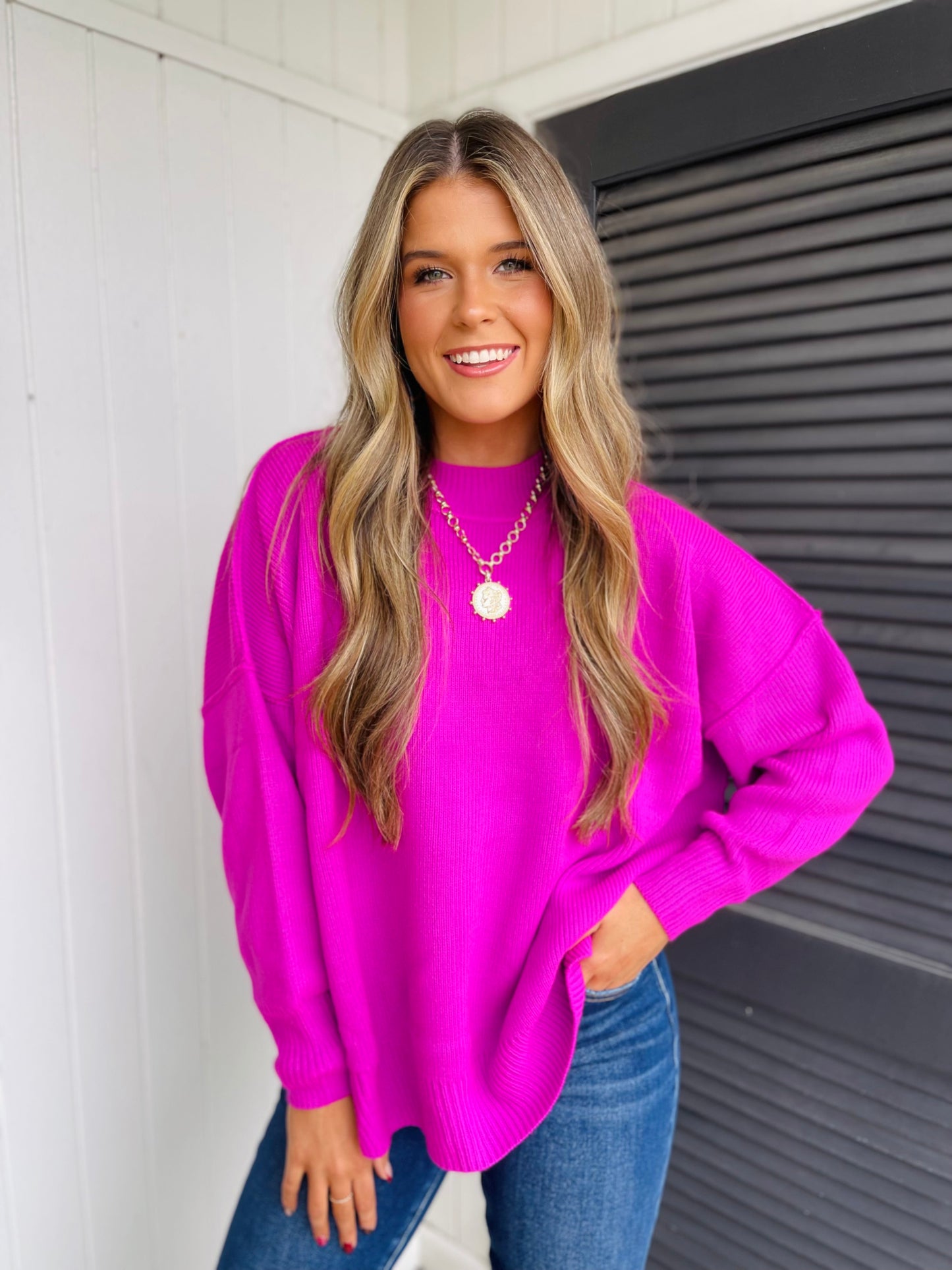 Woman wearing bright pink oversized sweater with gold necklace and blue jeans, smiling at camera.
