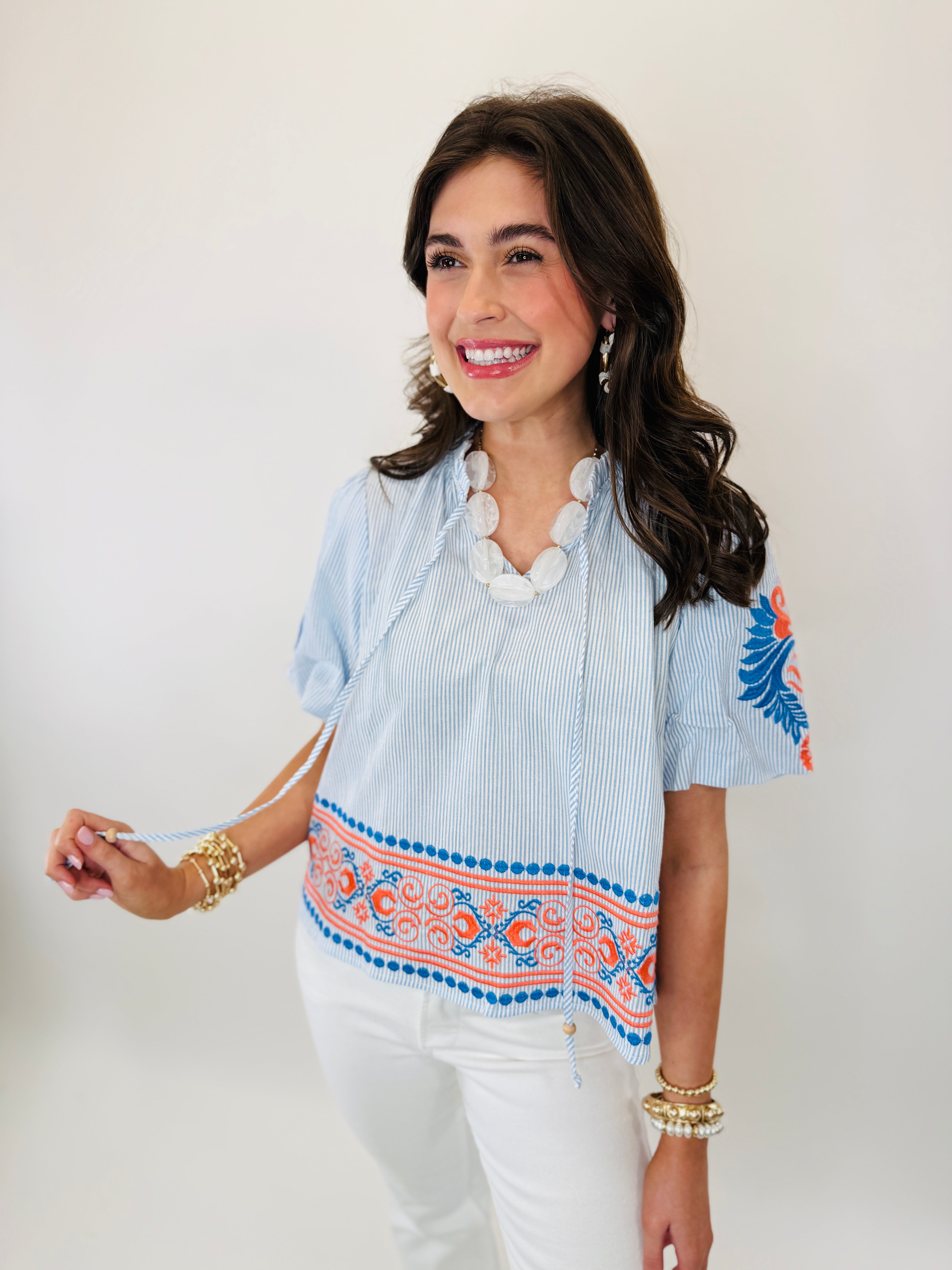 Woman wearing light blue striped Marcie top with colorful embroidered hem detail and white jeans.
