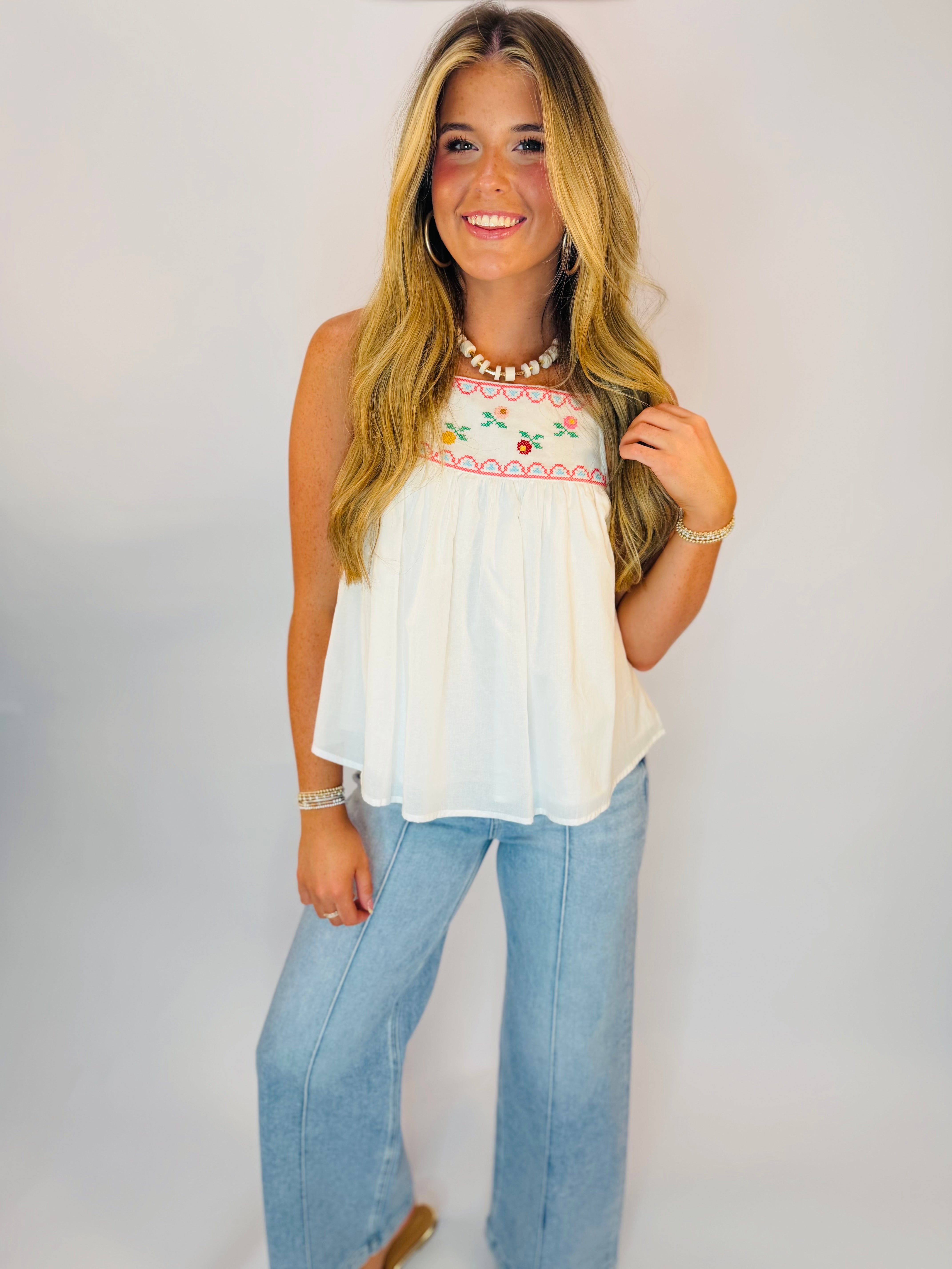 White sleeveless embroidered floral tank top with cross-stitch detail, styled with light wash wide-leg jeans.