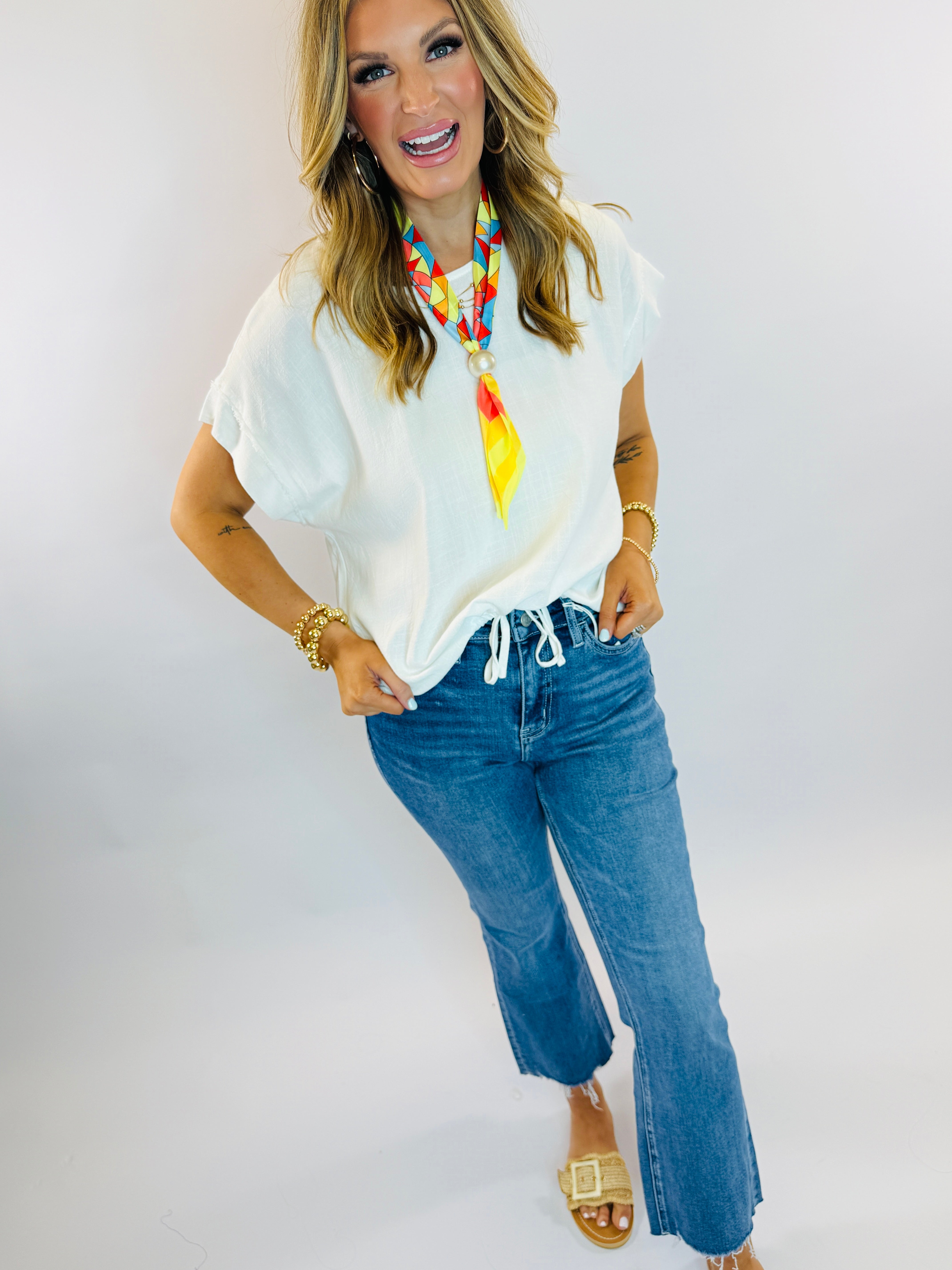 Woman wearing white linen top with colorful geometric scarf and blue jeans, styled with gold accessories.
