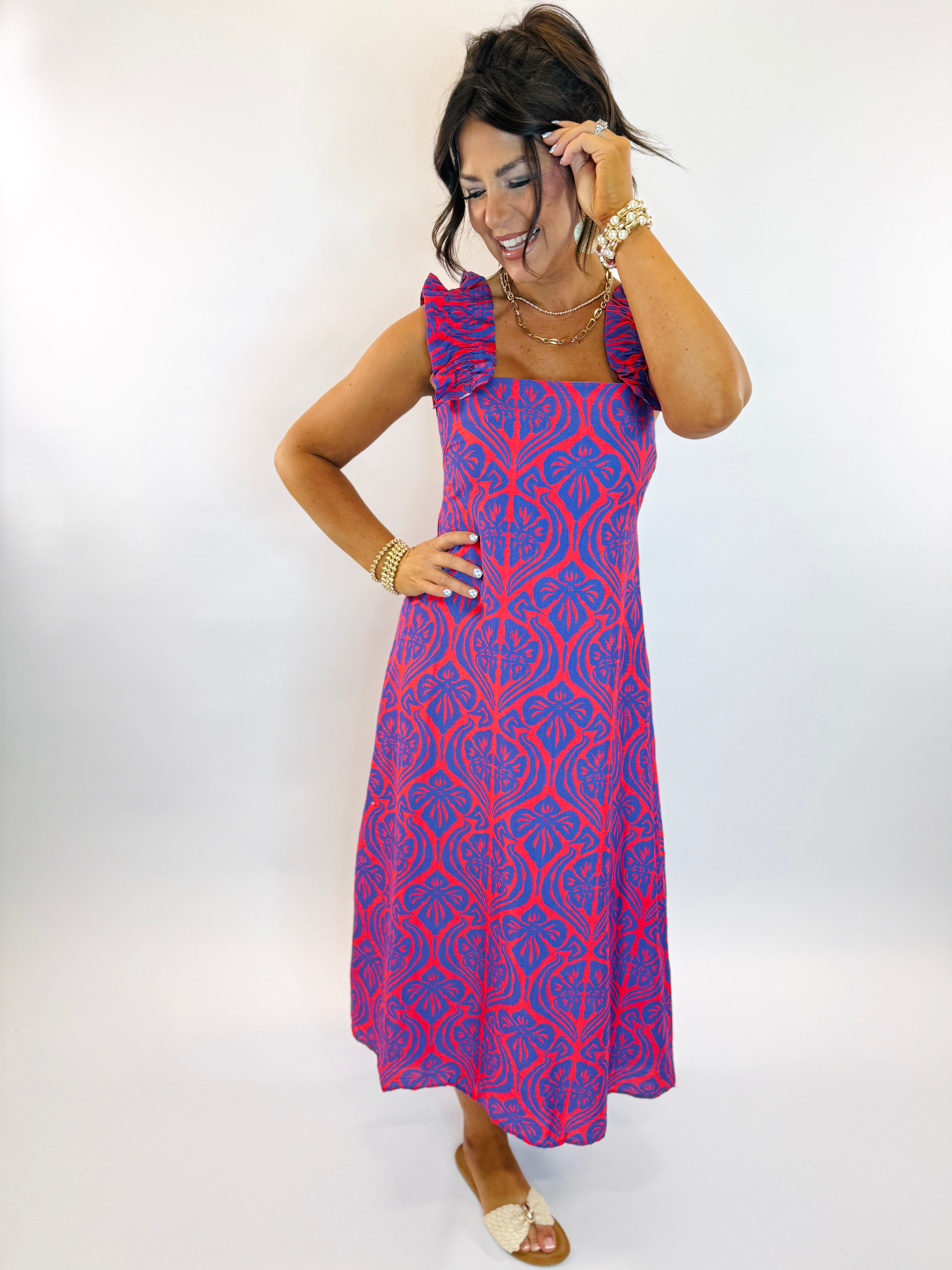 Woman wearing a red and blue patterned sleeveless midi dress with ruffle straps and gold jewelry accessories.