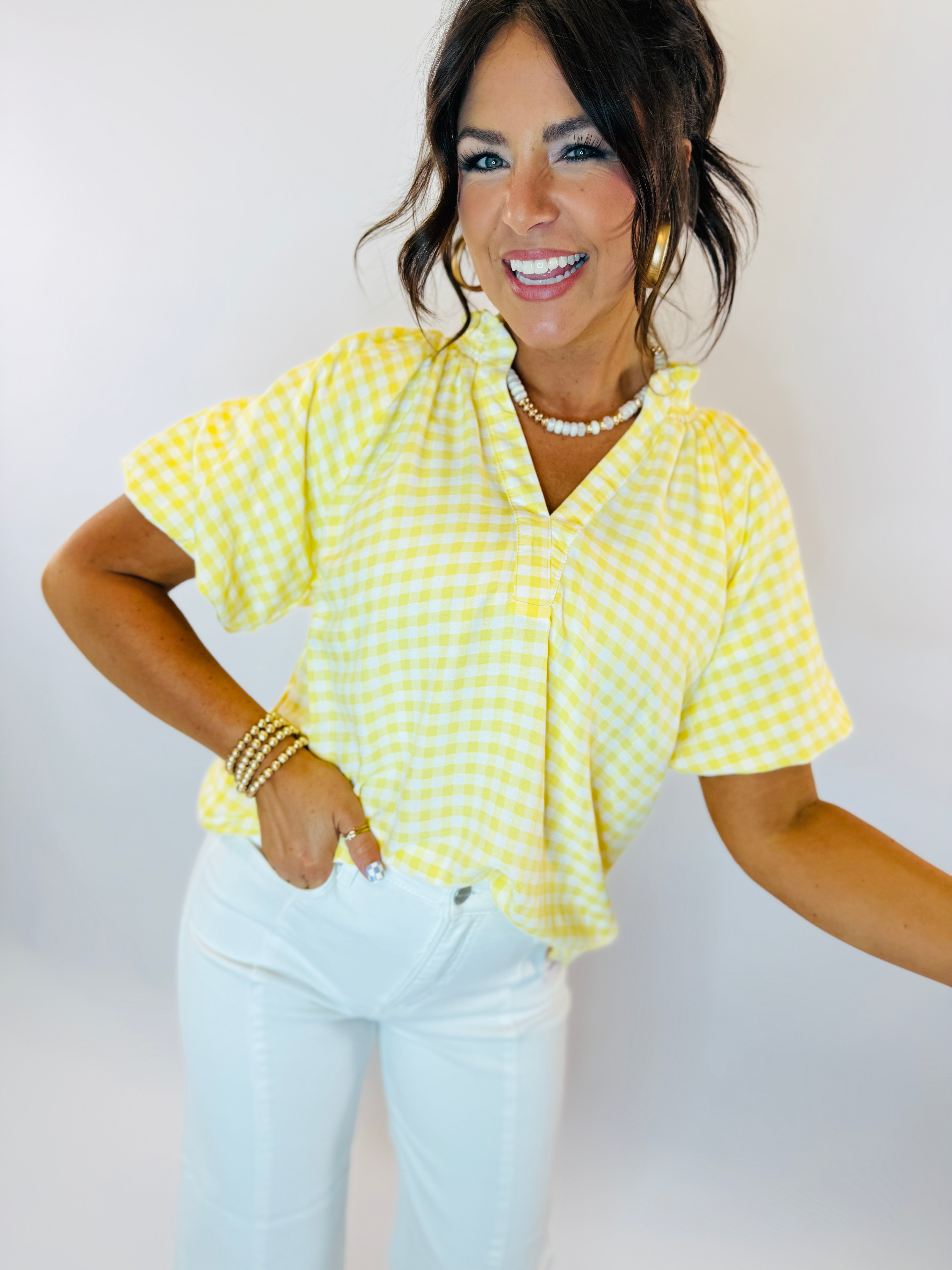 Woman wearing yellow and white gingham checkered short-sleeve button-up top with white jeans and pearl necklace.