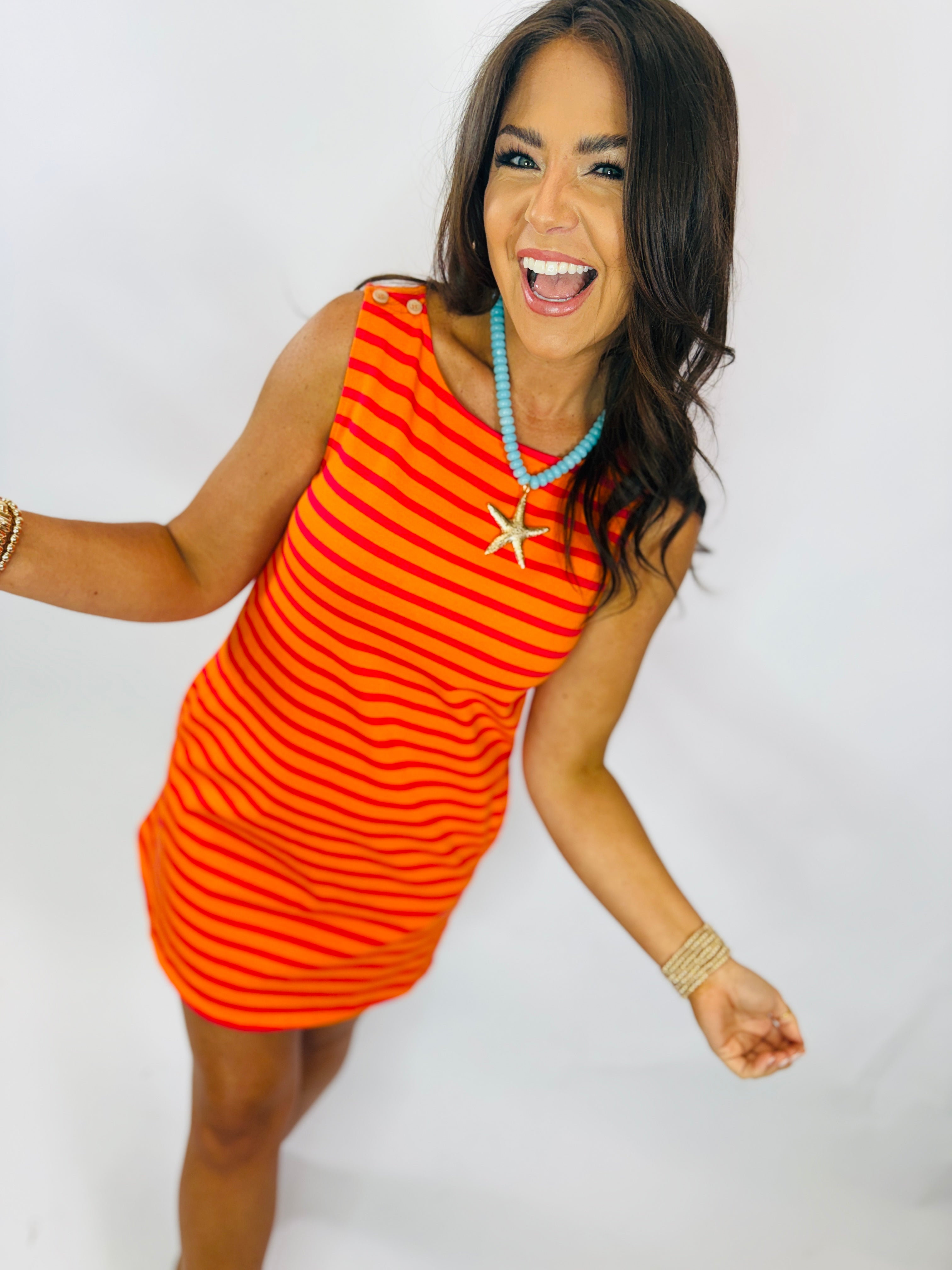Woman wearing orange and red striped sleeveless mini dress with turquoise necklace and gold bracelet, smiling against whit...
