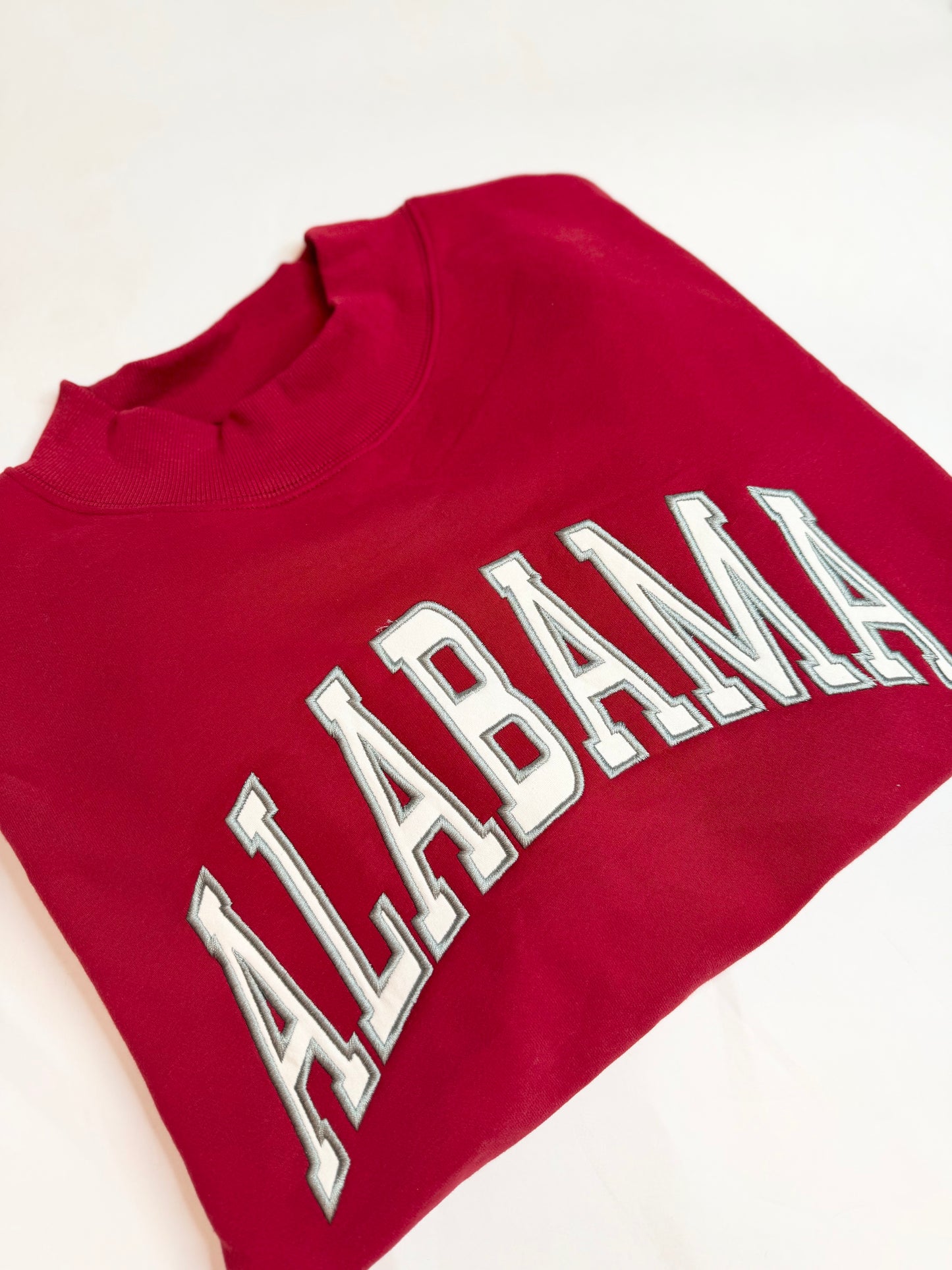 Crimson red mockneck sweatshirt with white embroidered appliqué "ALABAMA" lettering, folded flat.