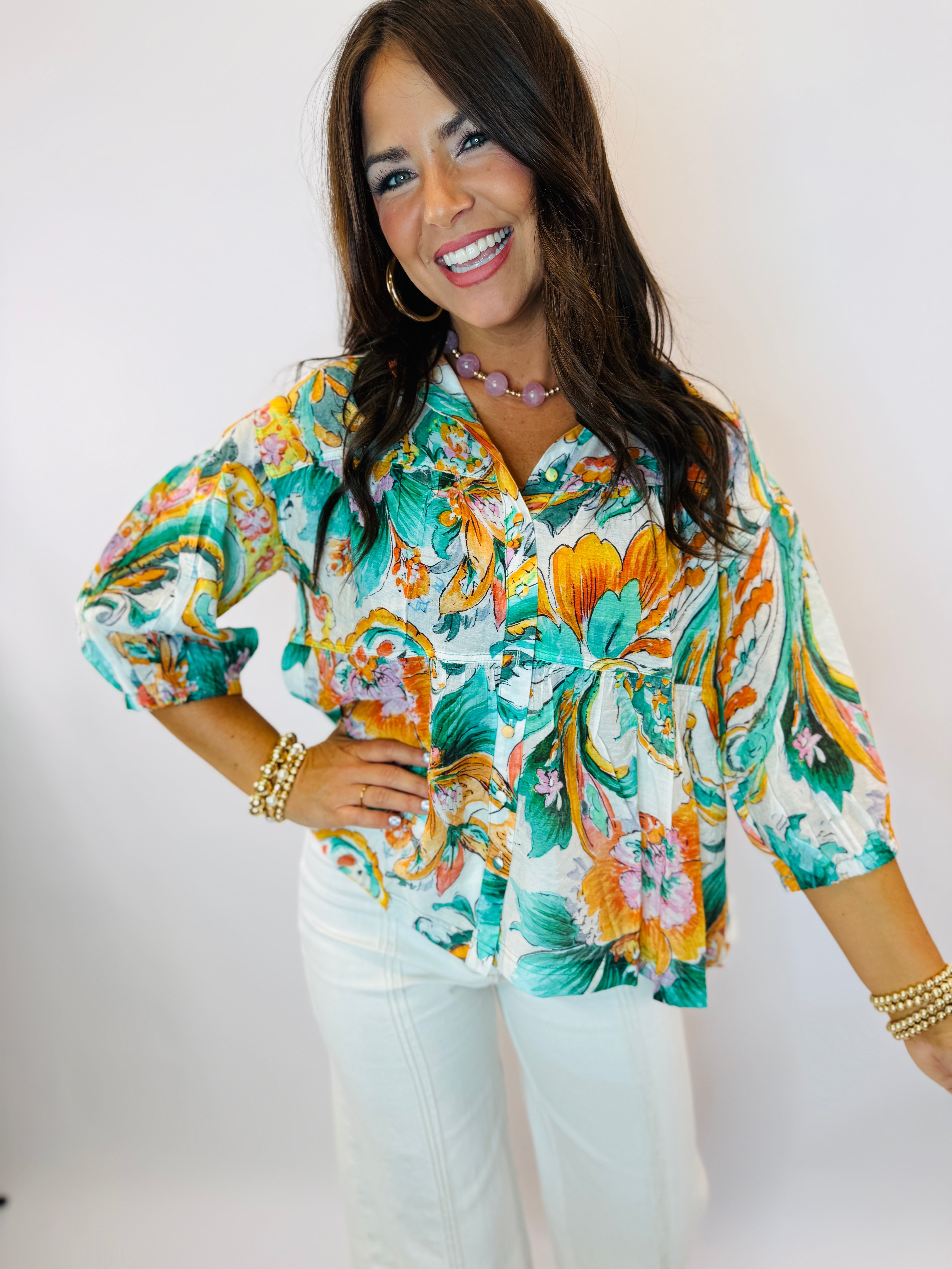 Woman wearing vibrant teal floral print blouse with three-quarter sleeves, styled with white pants and pearl necklace.