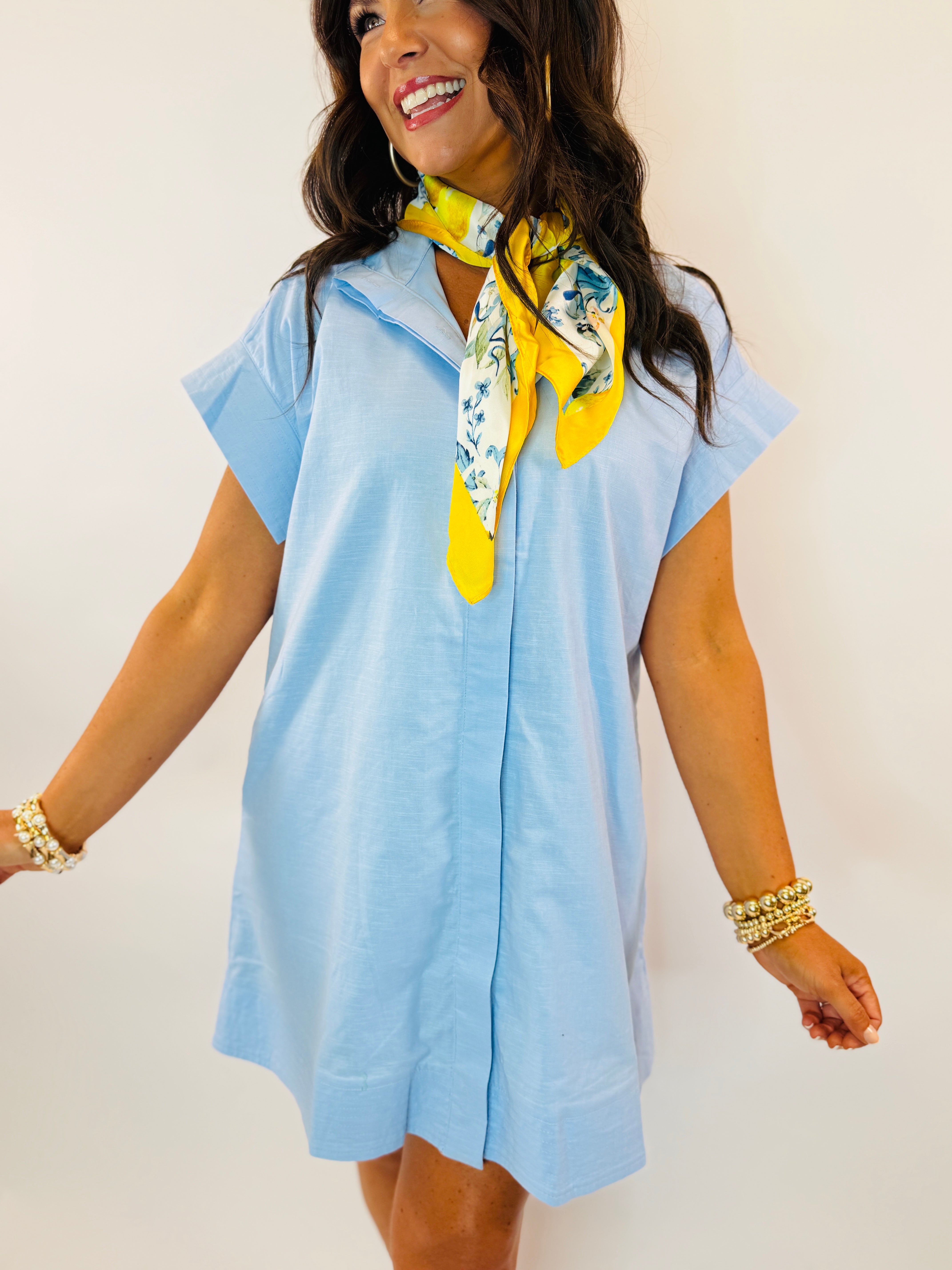 Woman wearing light blue short-sleeve Penelope dress with colorful floral scarf and pearl bracelets, smiling against white...