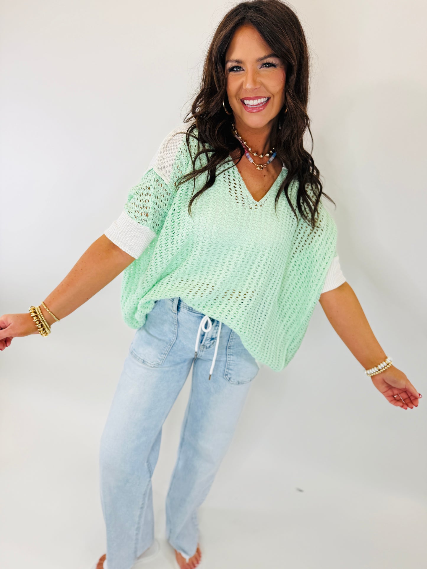 Woman wearing mint green knitted sweater with white contrast sleeves and light blue jeans, smiling at camera.