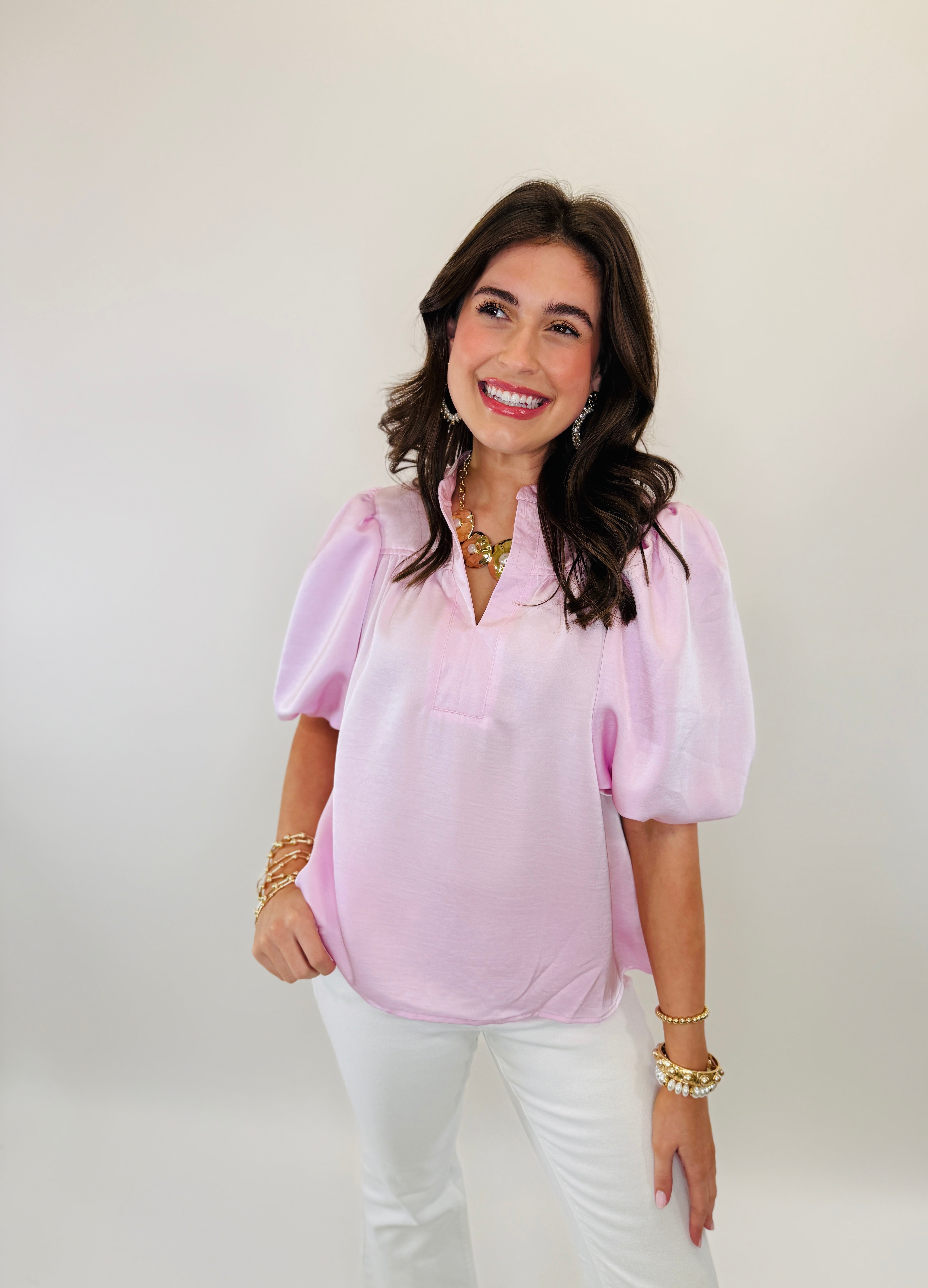 Woman wearing pink balloon sleeve blouse with white jeans and gold jewelry, smiling at camera