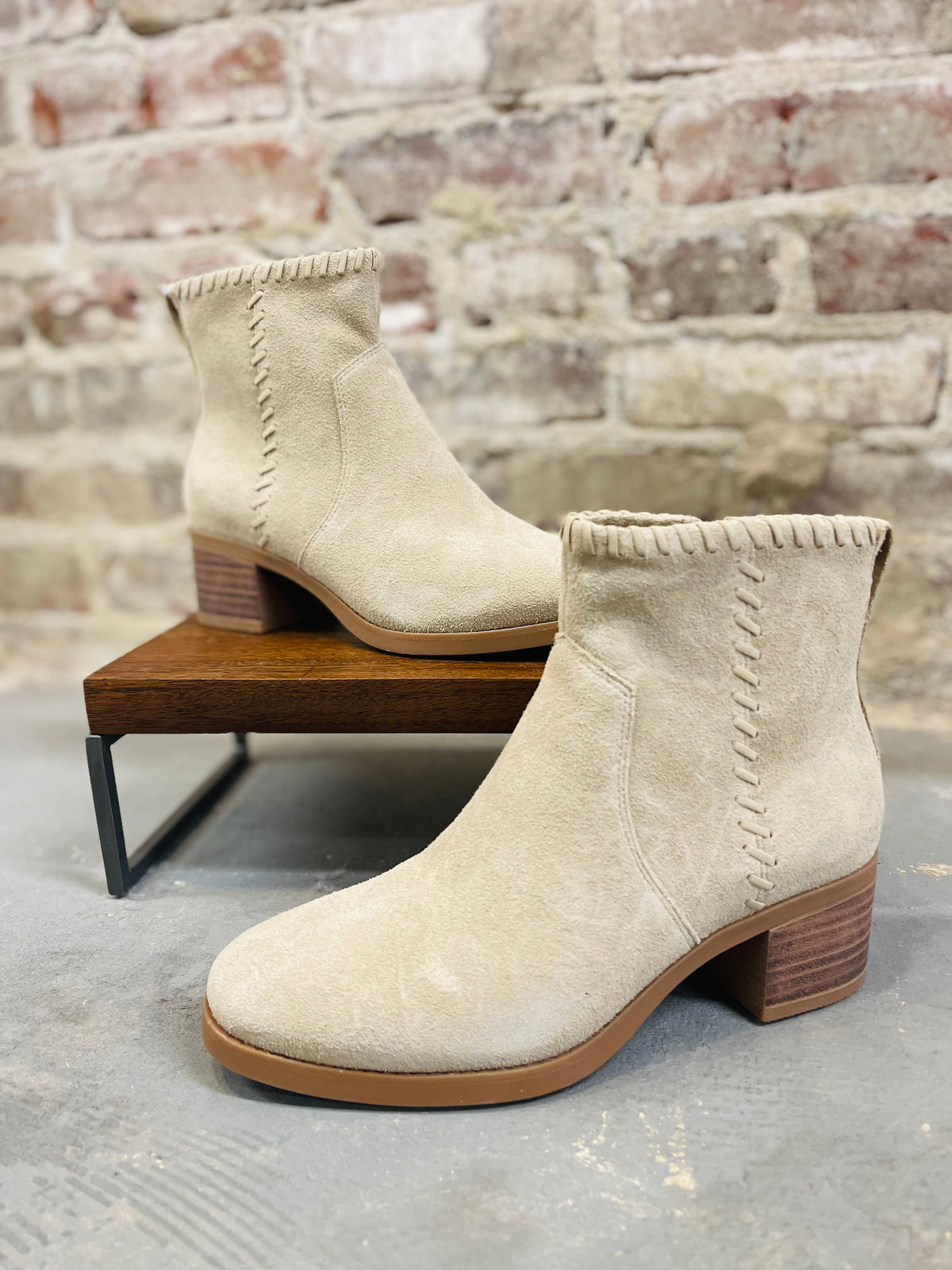 Beige suede Delilah ankle booties with decorative stitching and block heels displayed on wooden bench