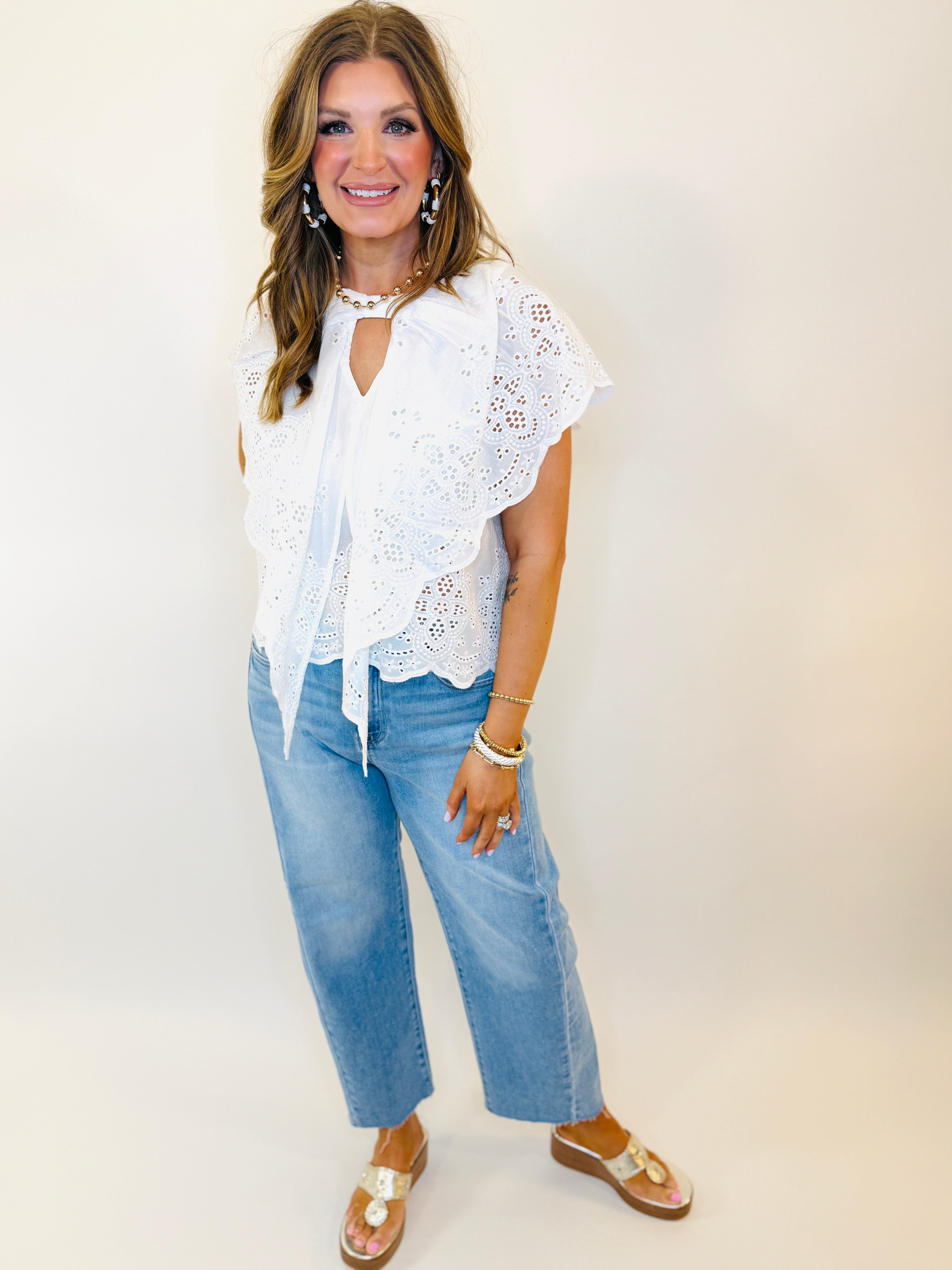 Woman wearing white eyelet lace top with tie front and blue wide-leg jeans, styled with sandals and jewelry.