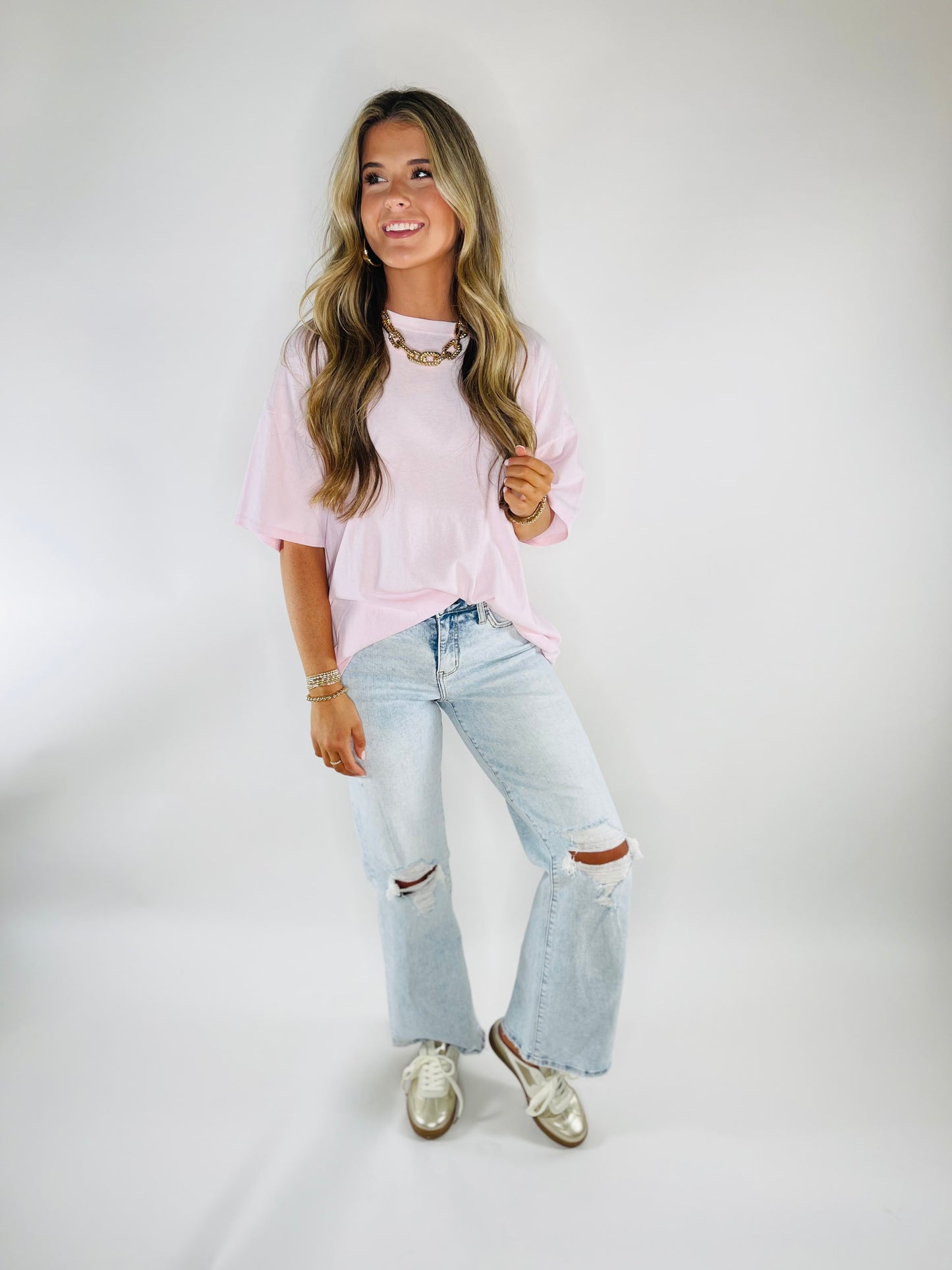 Woman wearing pink oversized Danna top with ripped jeans and gold jewelry, smiling against white background.
