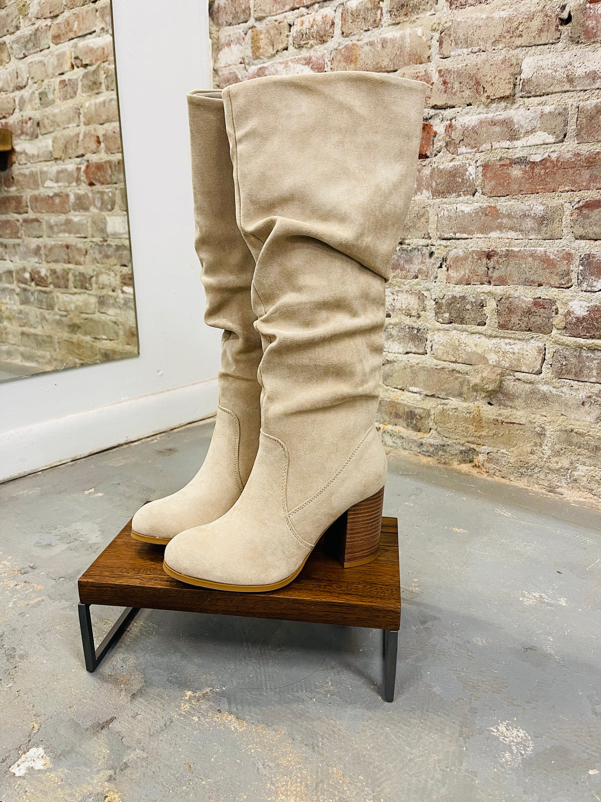 Beige suede slouchy knee-high boots with block heels displayed on wooden platform against exposed brick wall.