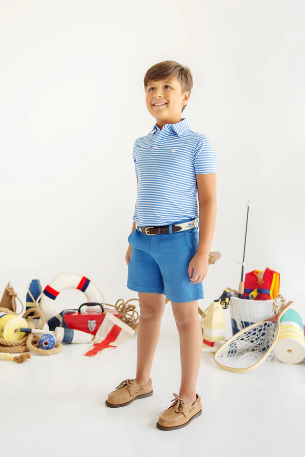 Young boy wearing blue and white striped polo shirt with blue shorts and boat shoes, standing among nautical toys.