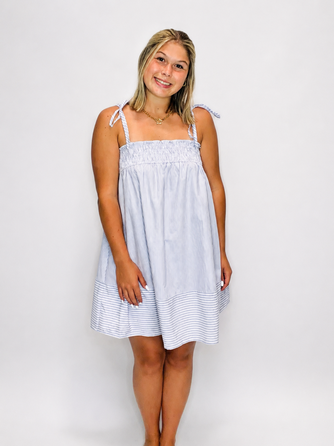 Young woman wearing light blue striped babydoll dress with tie shoulder straps and smocked bodice against white background.
